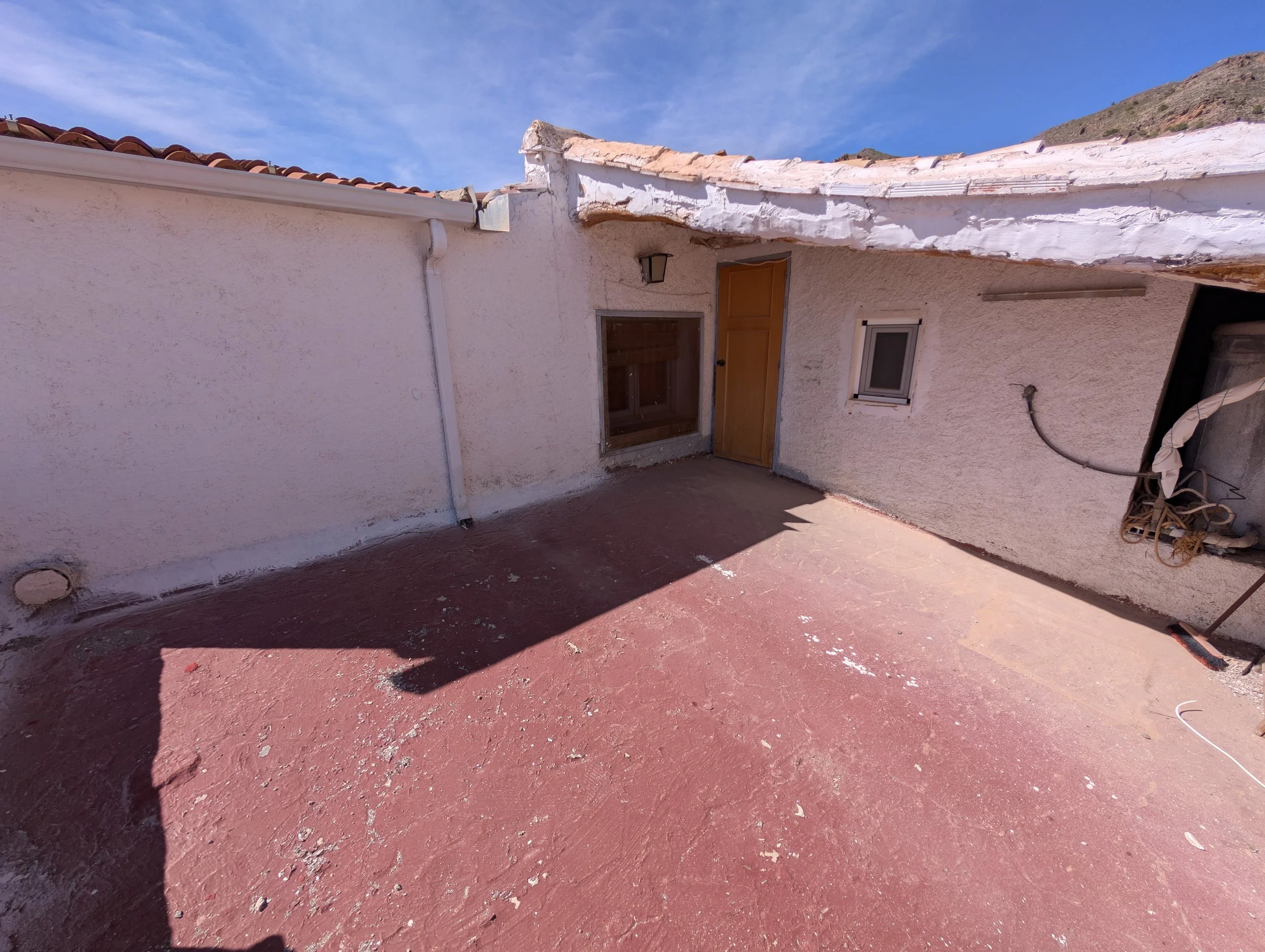 A small rooftop patio area with a textured pink concrete floor, beige stucco walls, a wooden door, and a small window, under a clear blue sky.