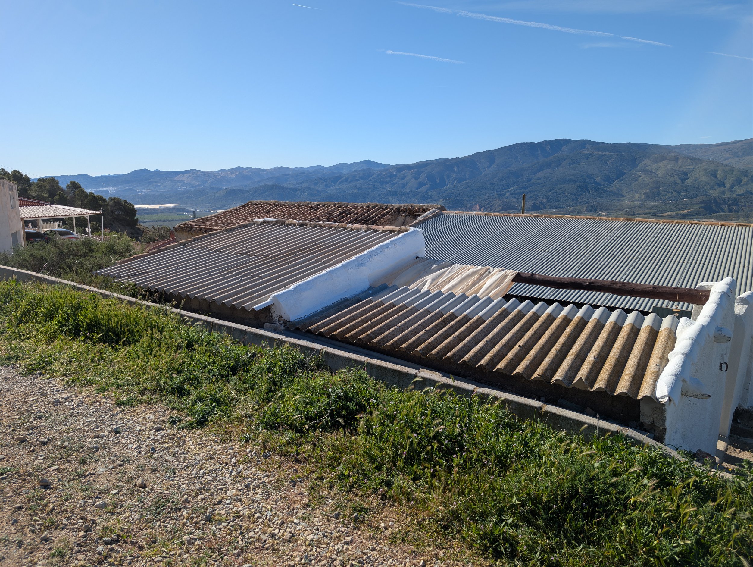 Rooftops made of corrugated metal and some with tiles, on a slope with greenery in foreground, mountains in background under clear blue sky.
