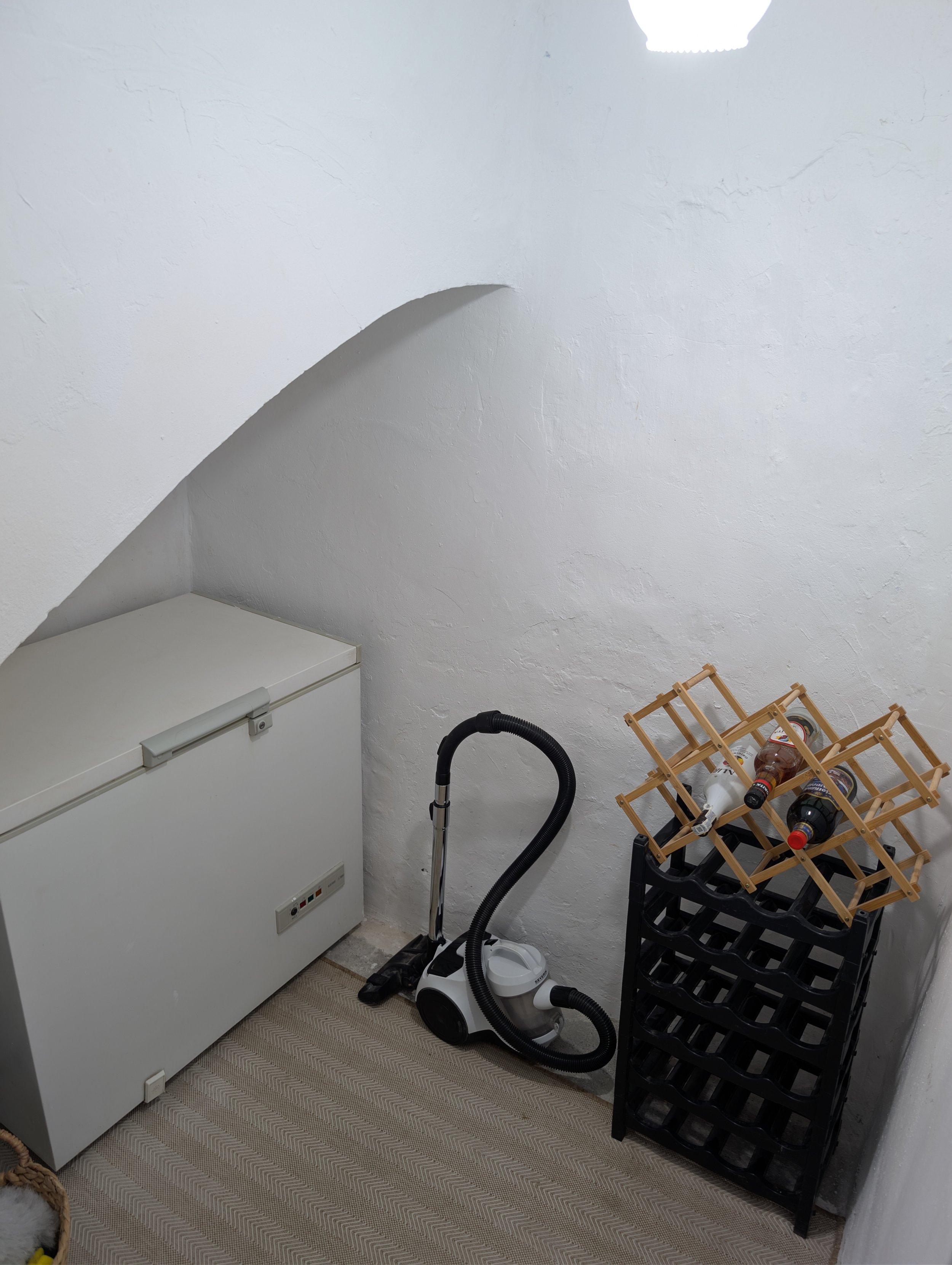 A small, cozy storage room with a white chest freezer, a vacuum cleaner, and a black wine rack filled with four bottles of soda or beer.