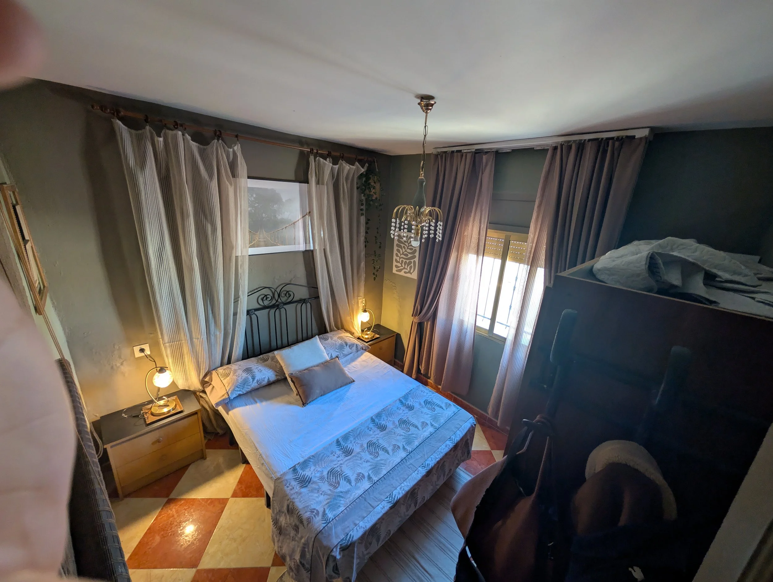 A bedroom with a double bed, bunk bed, nightstand, and curtains, illuminated by lamps and natural light from windows.