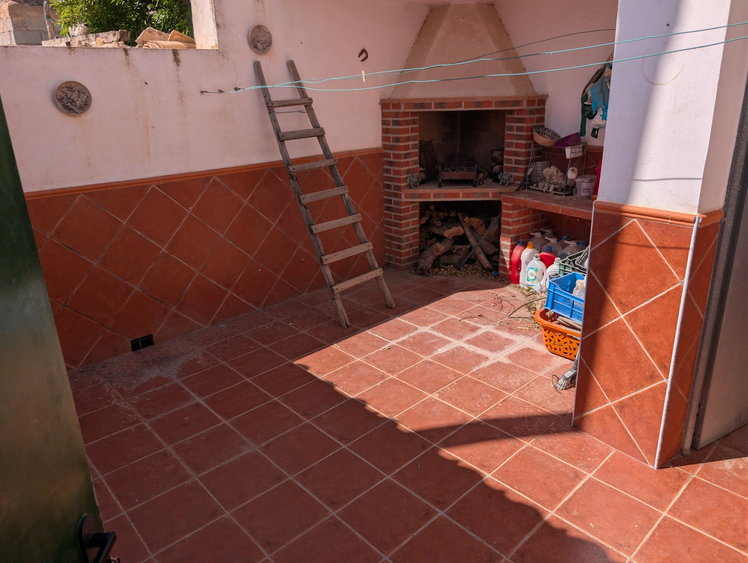 Outdoor patio with brick and tile fireplace, wooden ladder, and various cleaning supplies and gardening tools. Wall-mounted decorative plates and hanging dryer lines are also visible.
