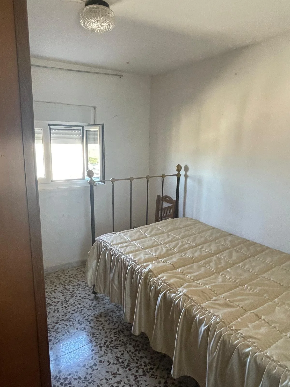 A small bedroom with a single bed covered in a beige quilt, a window with open shutters, a wooden chair next to the bed, a ceiling light fixture, and a terrazzo floor.