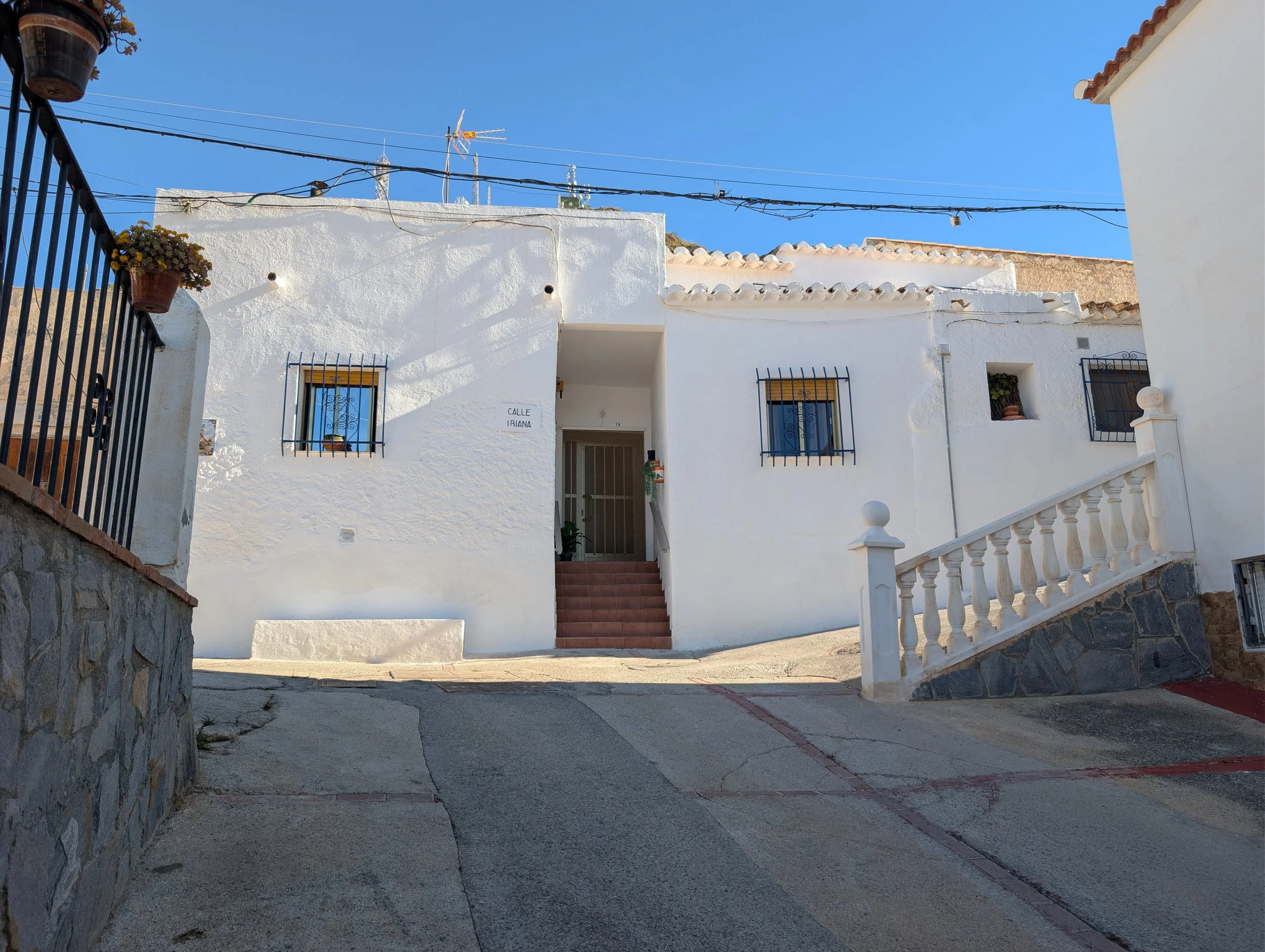 White stucco house with terracotta stairs leading to the entrance, black wrought-iron window bars, a staircase with a white railing on the right, potted plants, and a clear blue sky.