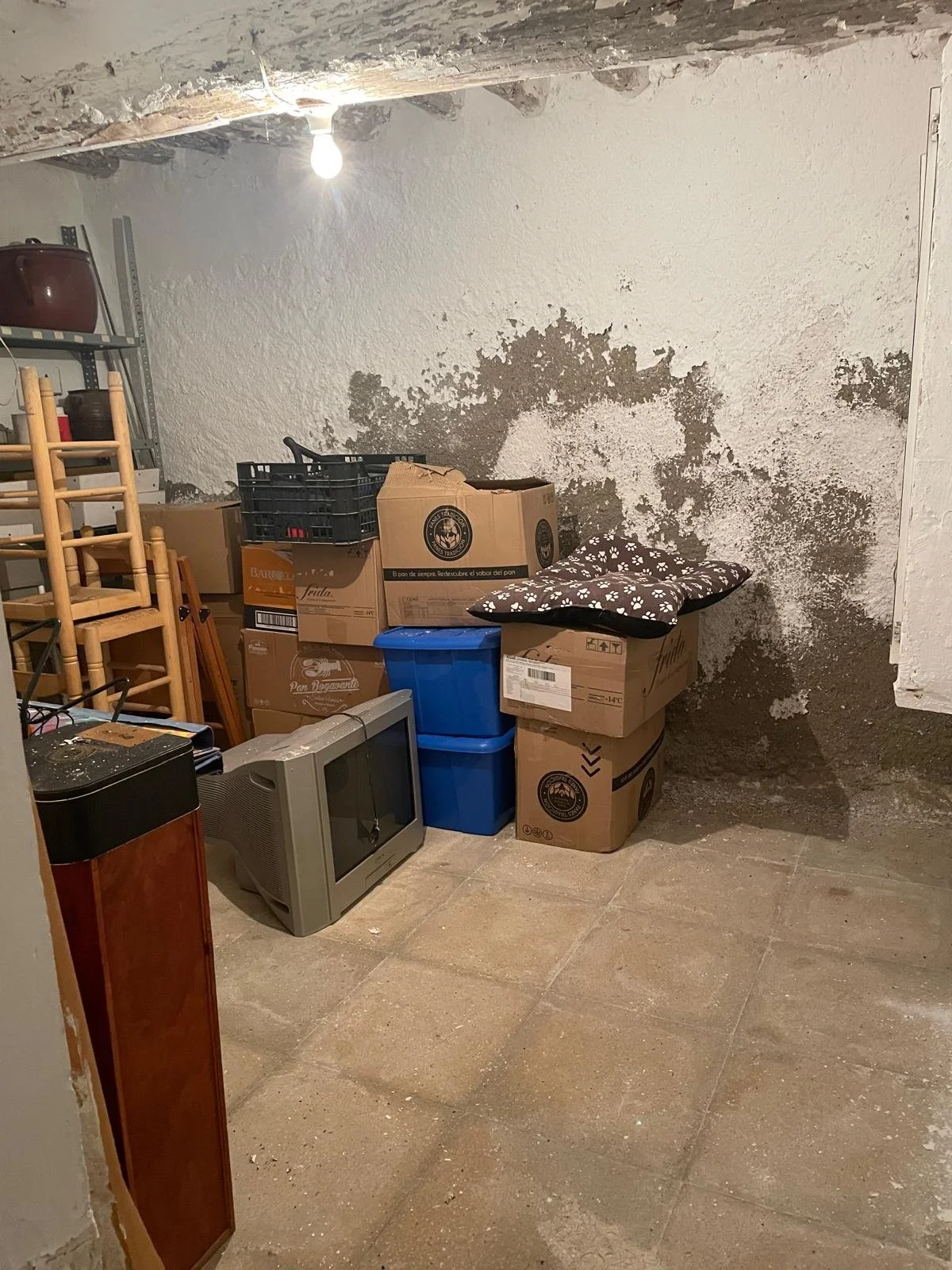A small storage room with peeling paint on the wall, cluttered with boxes, plastic containers, a small CRT television, and stacked chairs.