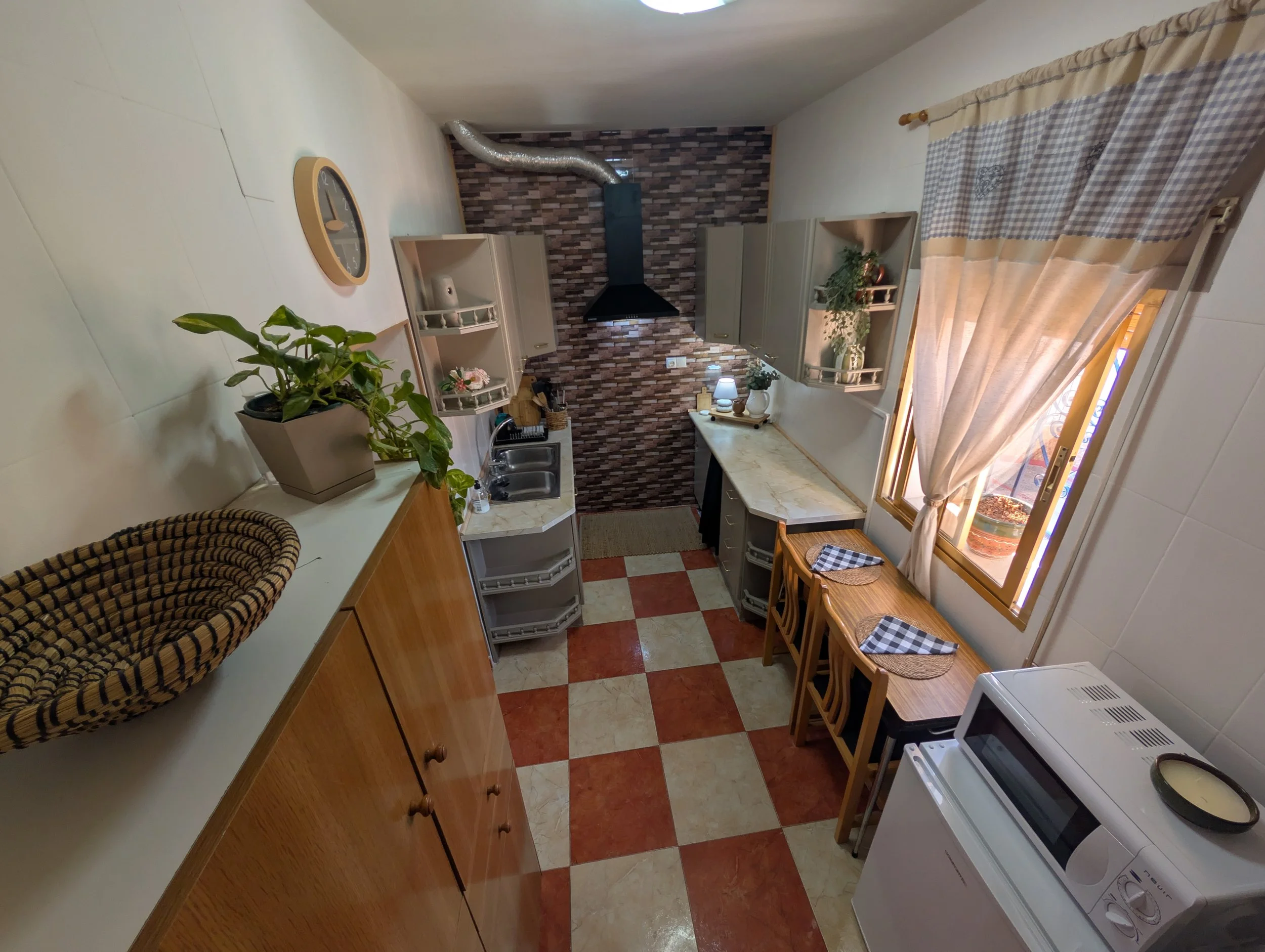 Small kitchen with tiled red and cream floor, window with beige curtains, wooden table with three chairs, kitchen counter with appliances, wall cabinets, and potted plants.