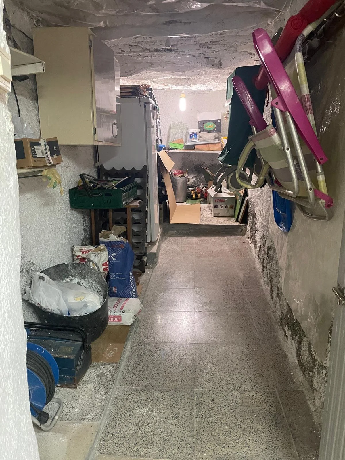 A cluttered basement storage room with various tools, boxes, and plastic chairs stacked against the wall.