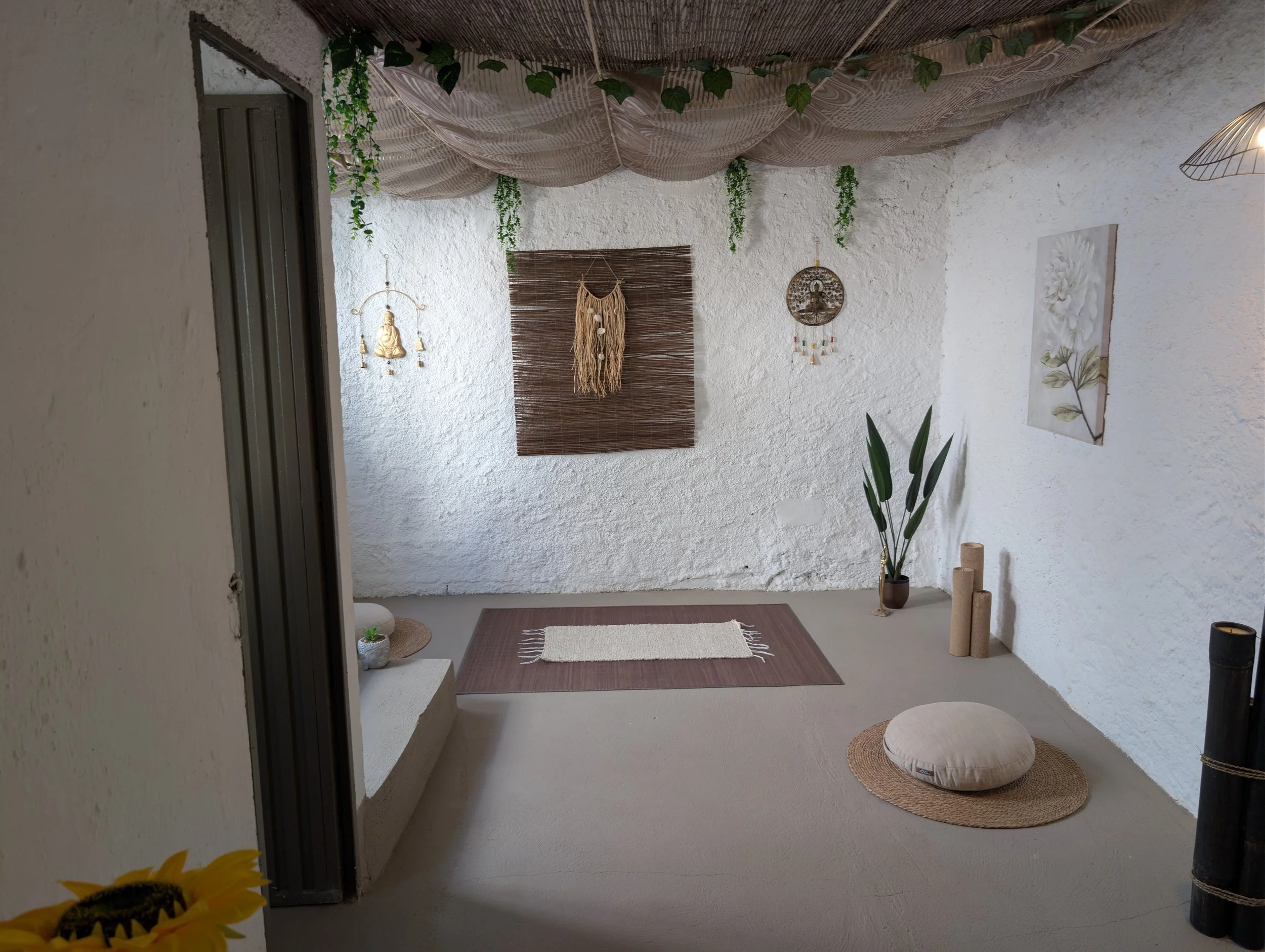 Bohemian-style meditation or relaxation space with white textured walls, green hanging plants, a wicker wall hanging, decorative wall art, a large plant in a pot, a beige cushion on a round woven mat, candles, a small area rug, and a bed of draped fa