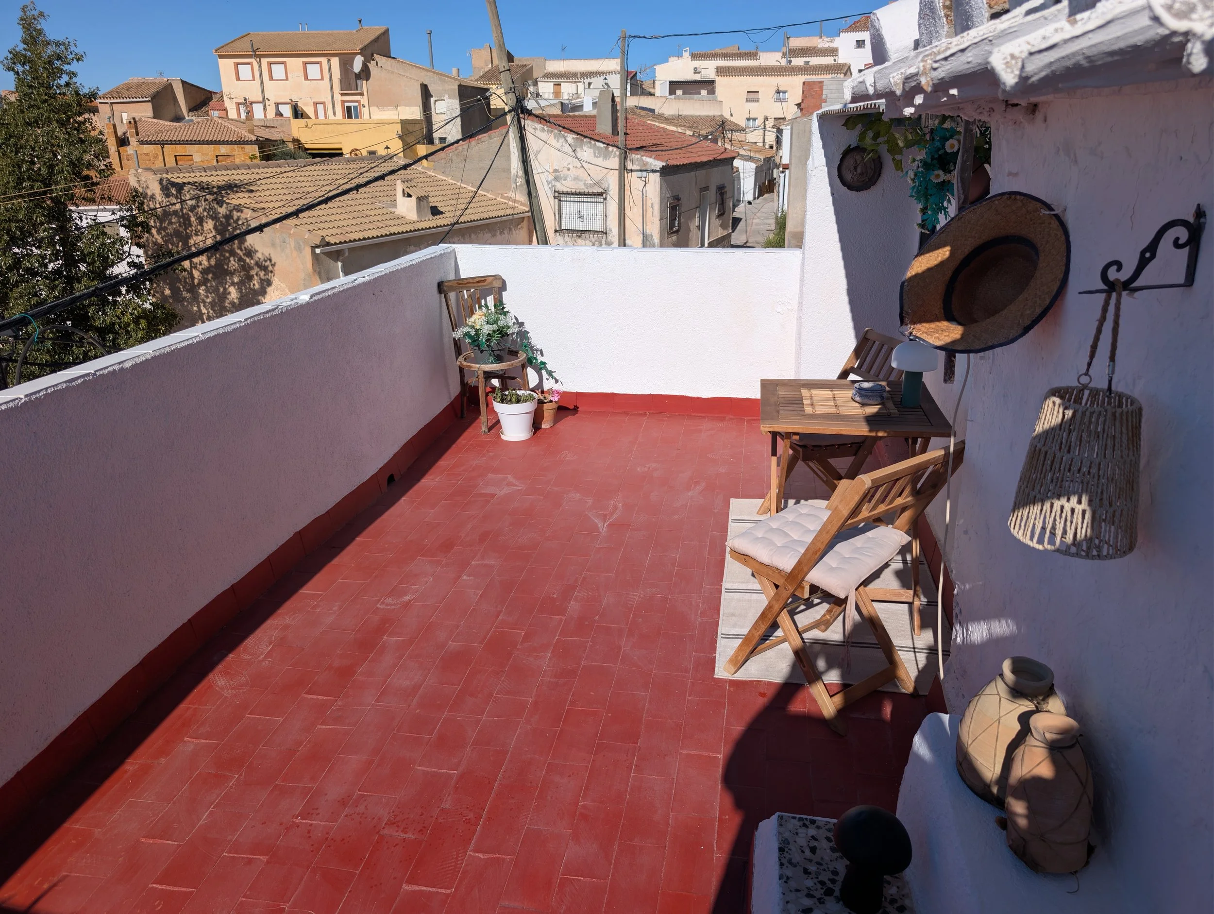 A rooftop terrace with red tile flooring, white walls, and outdoor wooden furniture including a small table and two chairs with white cushions. Decor items such as a hat, baskets, and potted plants are visible, with a view of neighboring buildings and a clear blue sky in the background.