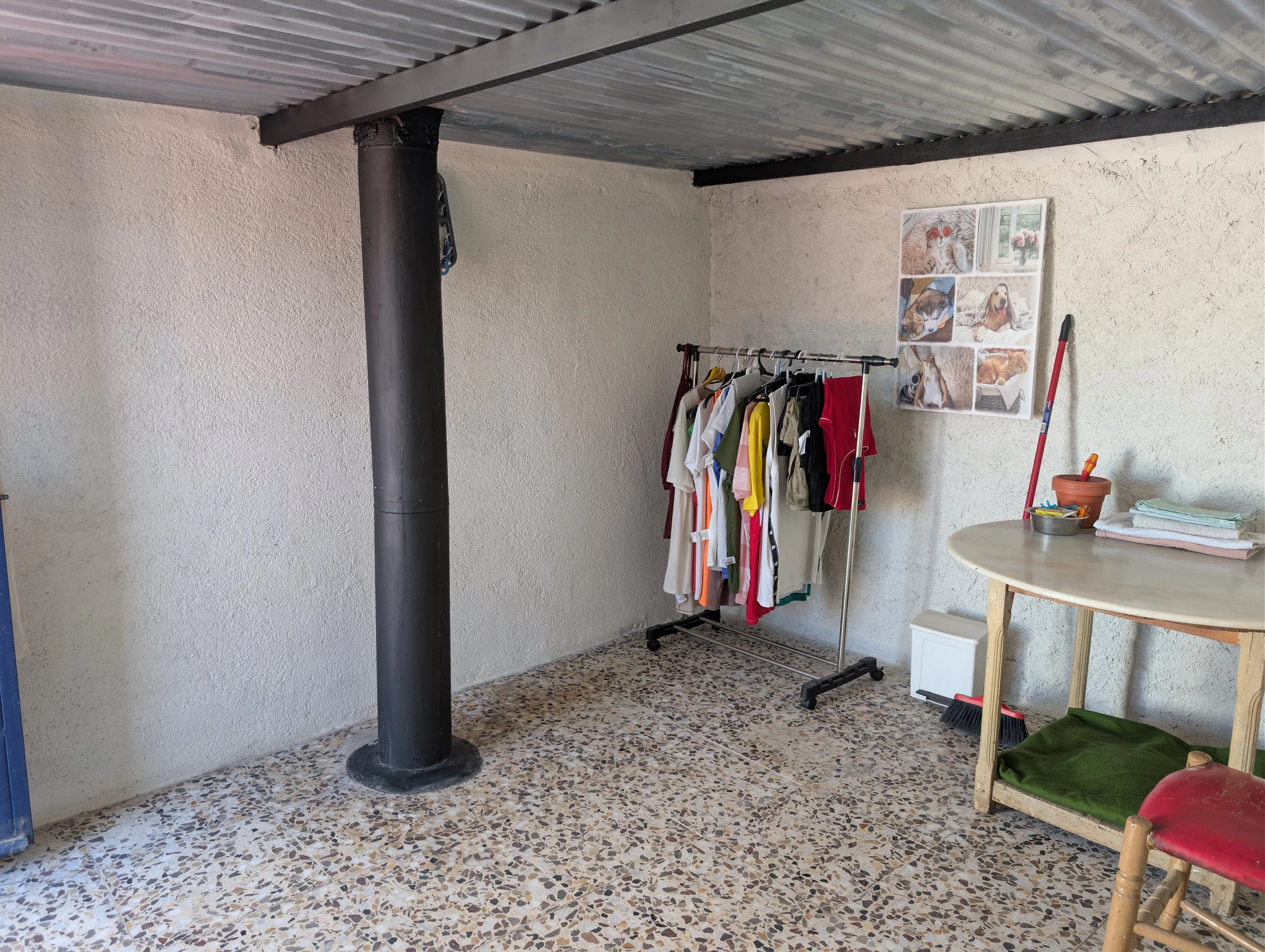 Room with clothes rack holding various shirts and pants, a round table with stacks of fabric or towels, a poster with dog photos on wall, cleaning tools leaning against wall, and terrazzo floor.