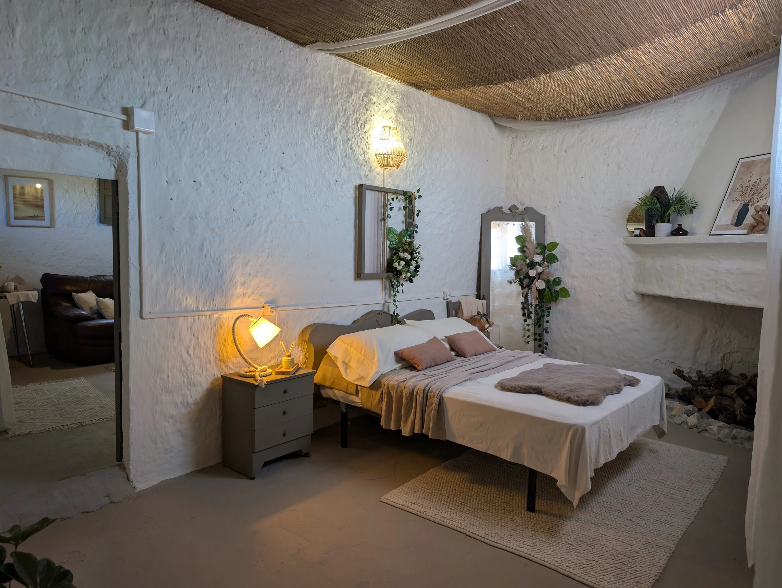 Cozy bedroom with white textured walls, a bed with white, beige, and pink bedding, decorative pillows, a gray nightstand with a lamp, a mirror with flowers, plants, and framed art, and an open doorway leading to a living area with a leather sofa.