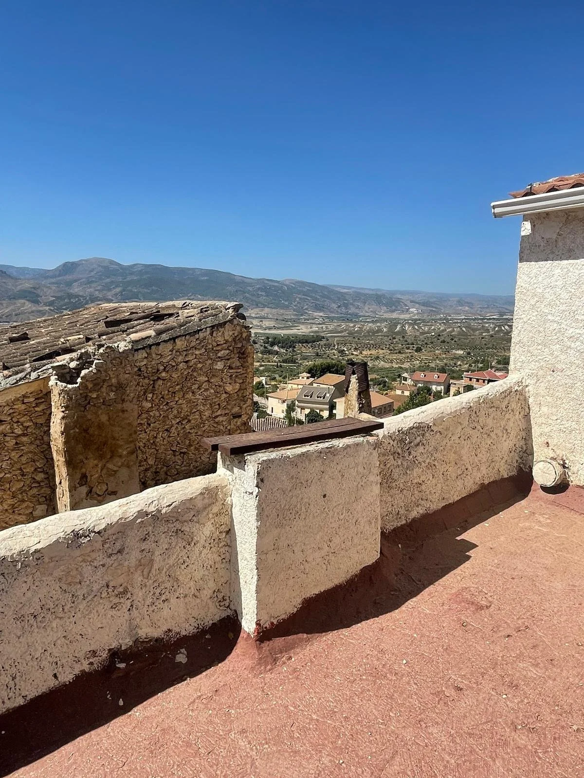 A view from a rooftop balcony with a stone and stucco wall, overlooking a small town and mountains under a blue sky.