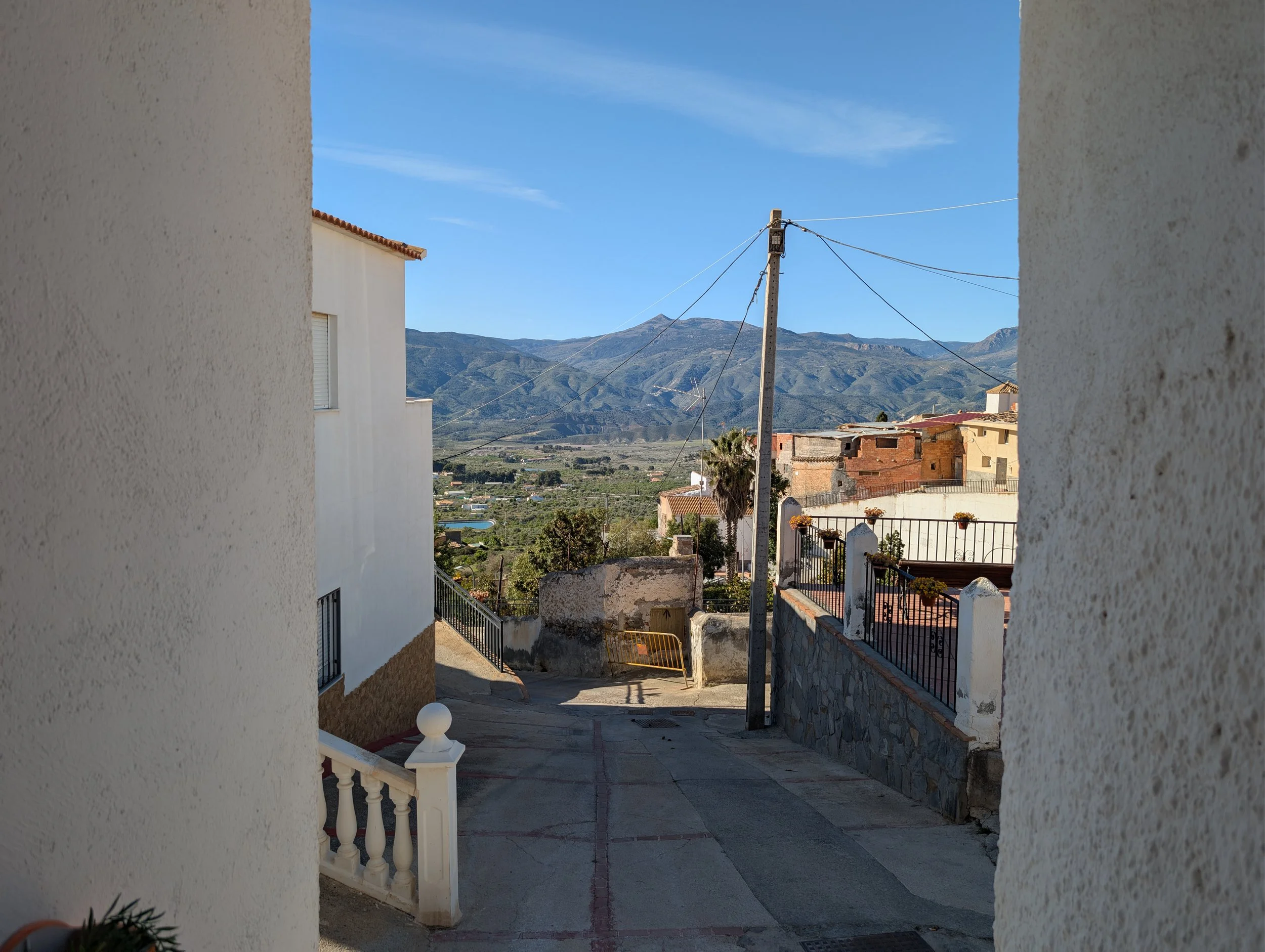 A narrow street view in a mountainous town, with white residential buildings, a utility pole with wires, a stone wall, a palm tree, and a scenic mountain range in the background under a clear blue sky.
