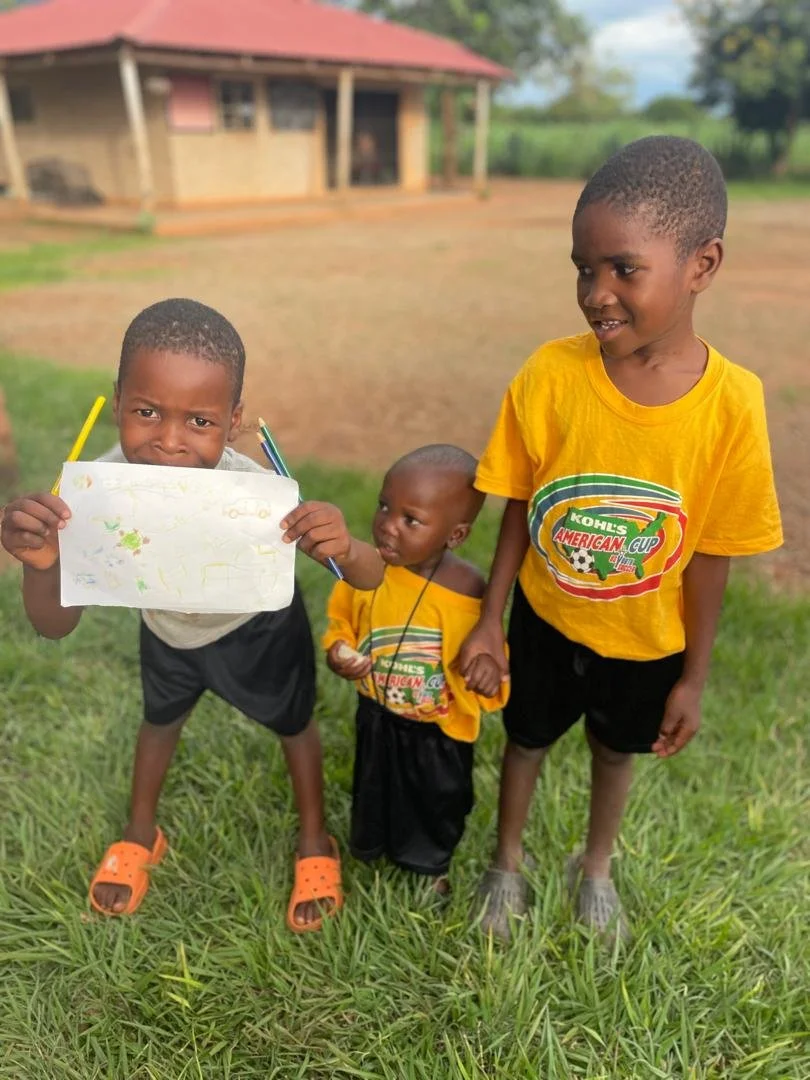 Three young boys standing on grass outdoors, with a house in the background. The boy on the left is holding a drawing and has a serious expression, wearing orange slip-on shoes. The boy in the middle is smaller, looking at the camera, wearing a yellow T-shirt with a soccer-themed logo and black shorts. The boy on the right, taller, is smiling and wearing a yellow T-shirt with the same logo and black shorts.
