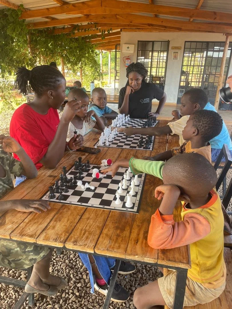 Children and an adult playing chess at a wooden table outside under a covered porch.
