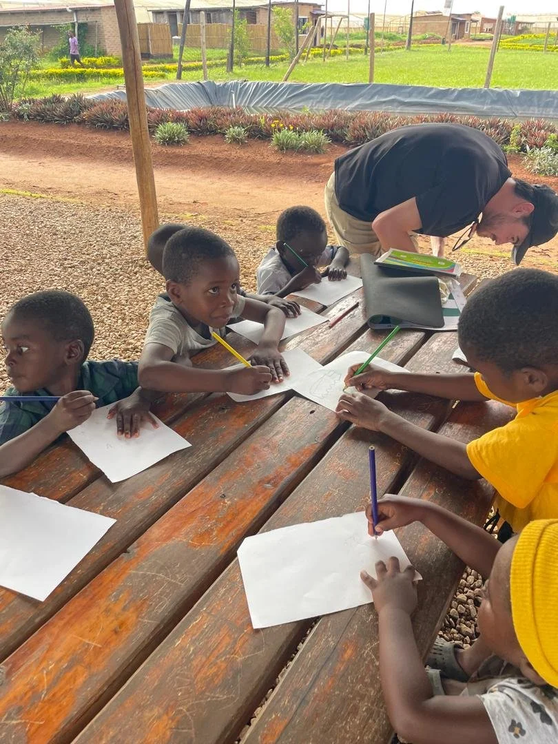 Children sitting at a wooden table outdoors, drawing on white paper with colored pencils, while an adult assists them. The setting appears to be a garden or farm with plants and a fence in the background.