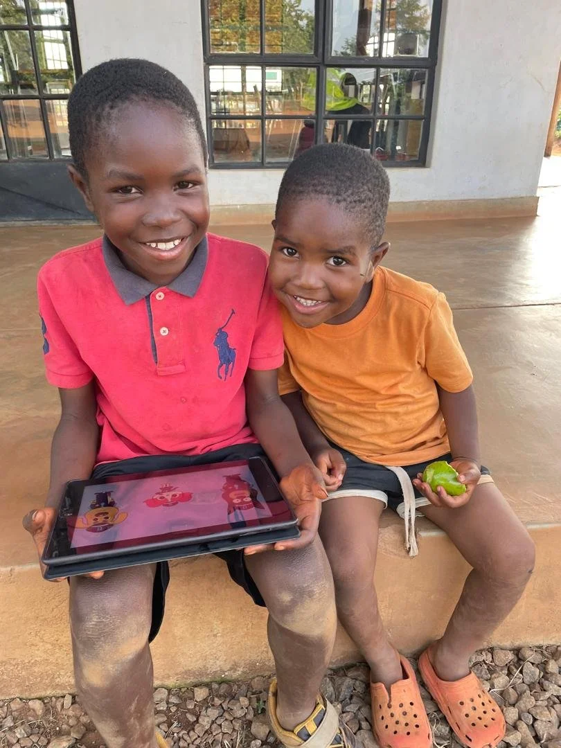 Two young boys are sitting on a concrete ledge. One boy, wearing a red polo shirt, is holding a tablet and smiling. The other boy, in an orange shirt, is holding a green pepper and also smiling. They appear to be outdoors, with buildings and windows behind them.