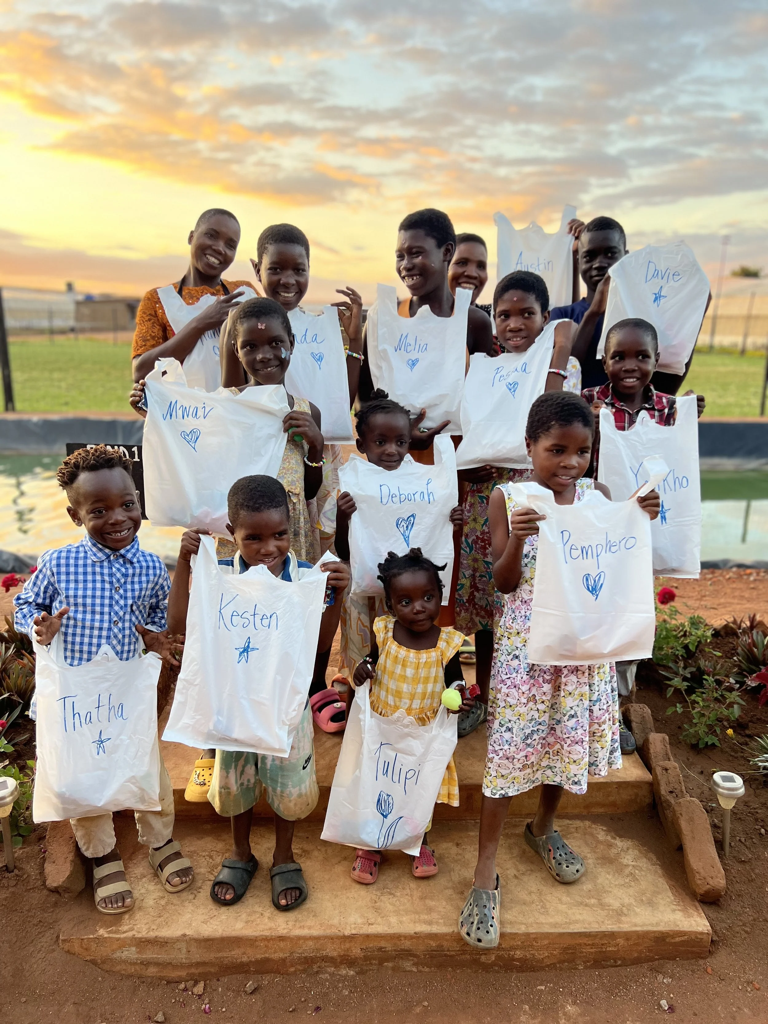 Group of children and adults outdoors at sunset, holding paper bags with handwritten names and drawings, standing on a brick platform with a garden and water feature in the background.