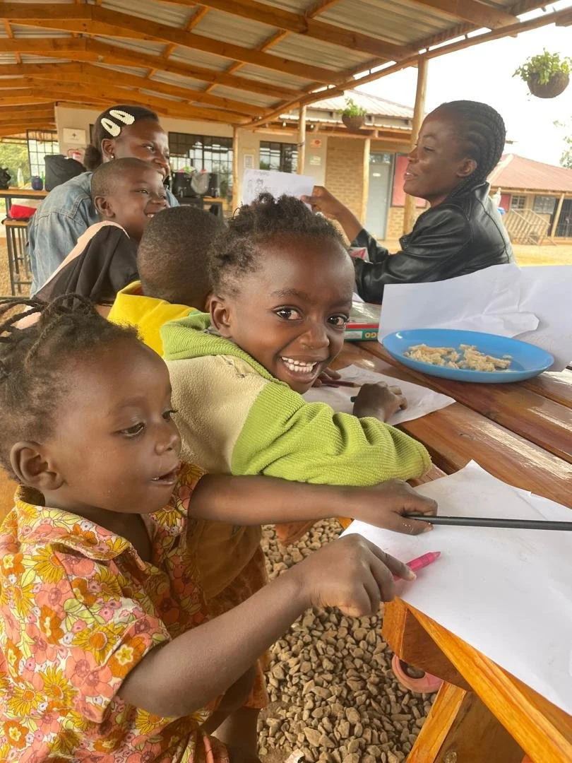 Children and adults sitting at a wooden table under a roof, talking and smiling, with children holding pens and paper.