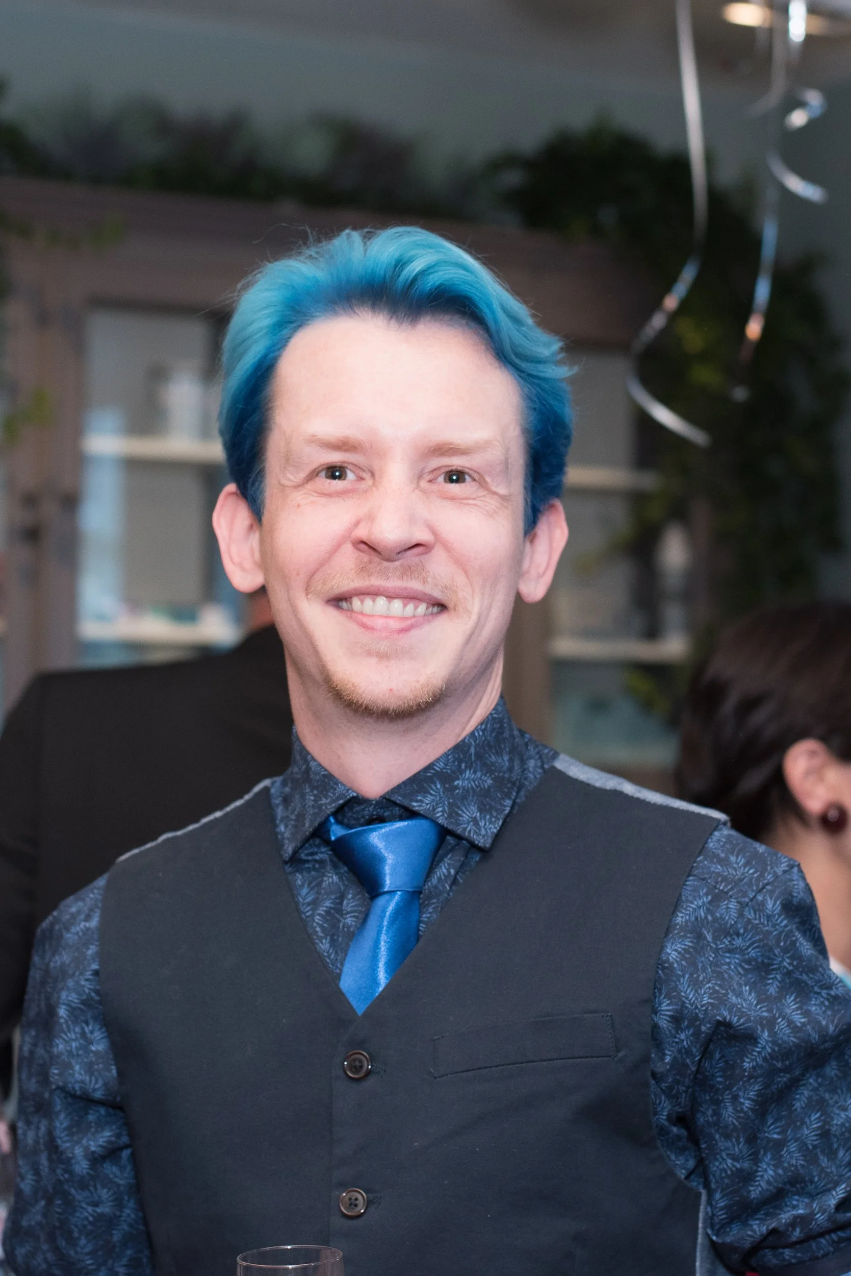 Portret van een man met blauw haar in een gilet, glimlachend tijdens een evenement.
