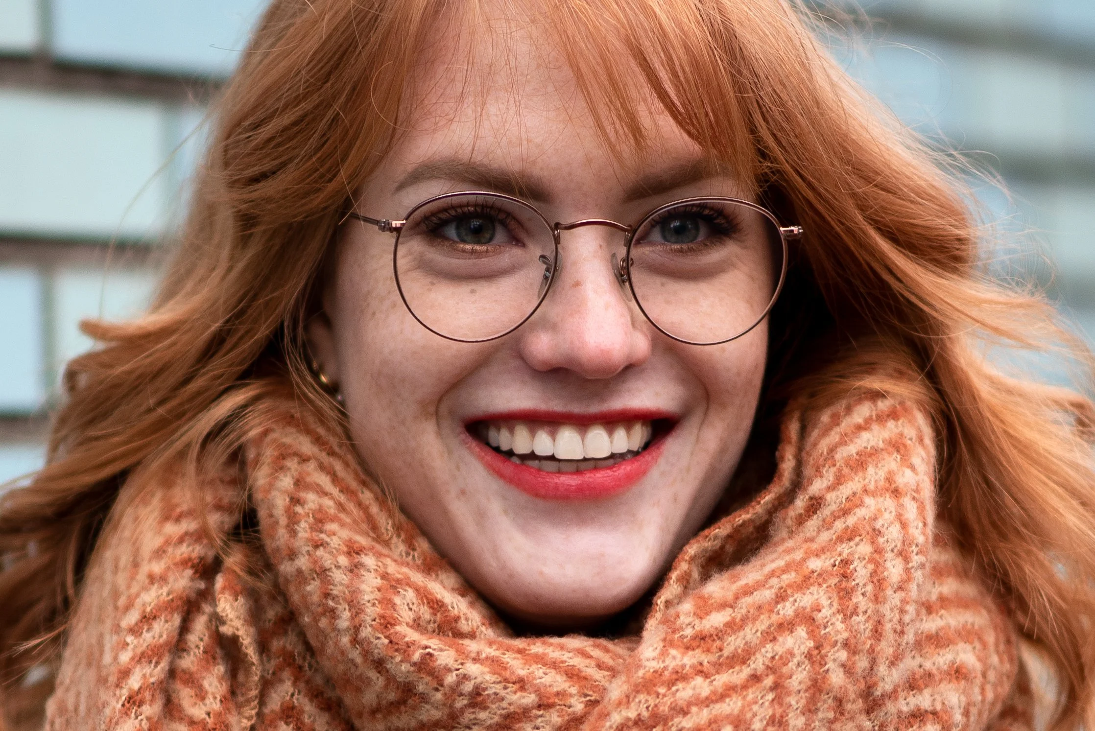 Portret van een lachende roodharige vrouw met een ronde bril en een warme sjaal.