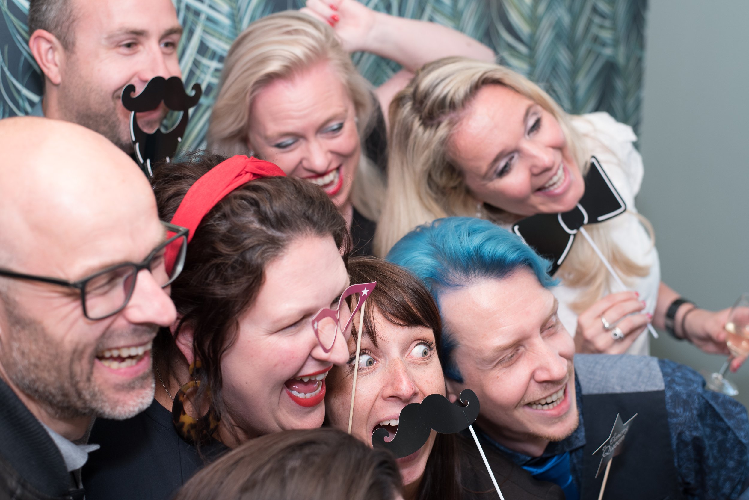 Gezellige groepsfoto van gasten met snorren op een stokje in een fotobooth tijdens een feest.