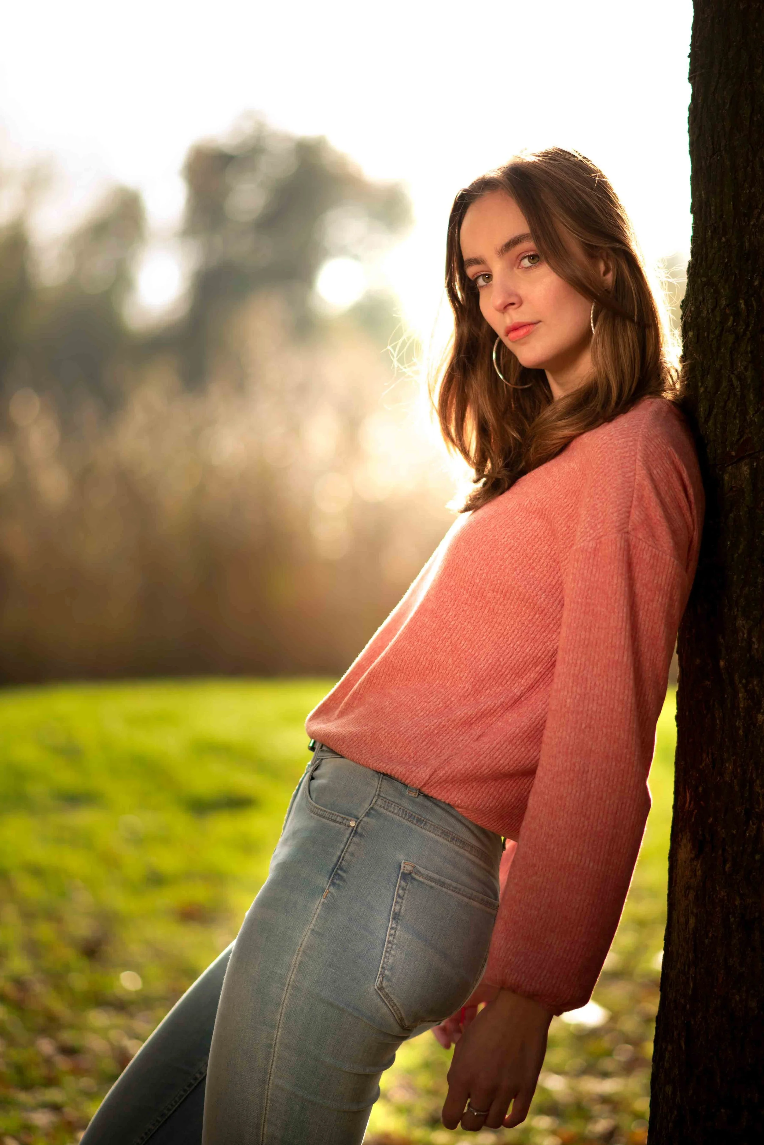 Romantisch portret in tegenlicht van een vrouw die leunt tegen een boom in een park.