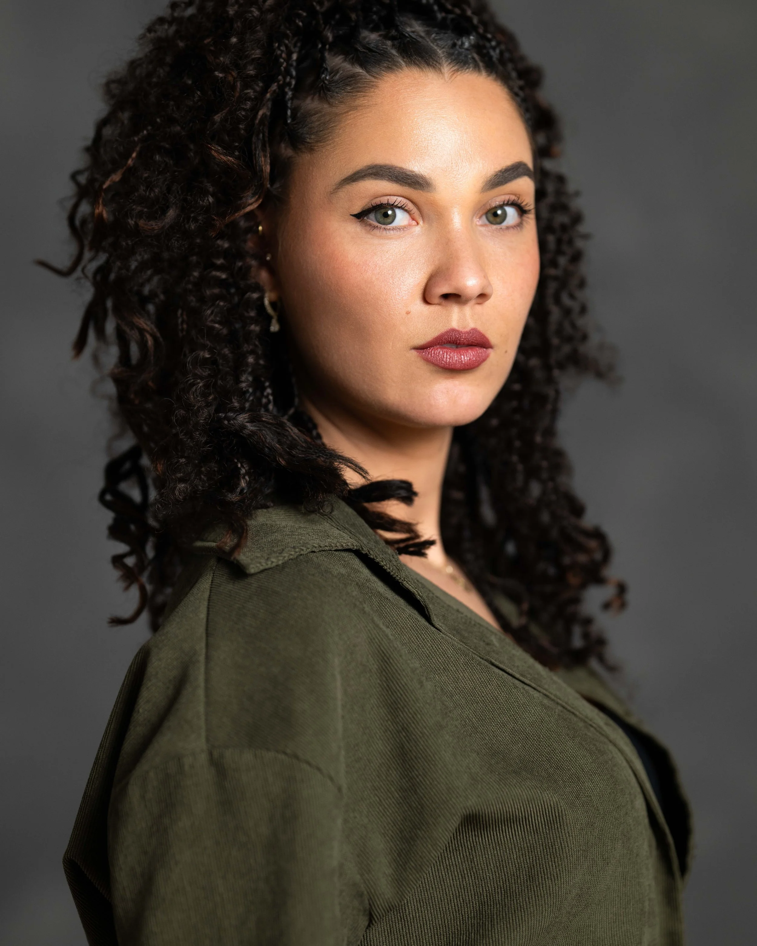 Professioneel studioportret van een jonge vrouw met krullend haar in een groene jas, gefotografeerd tegen een grijze achtergrond.