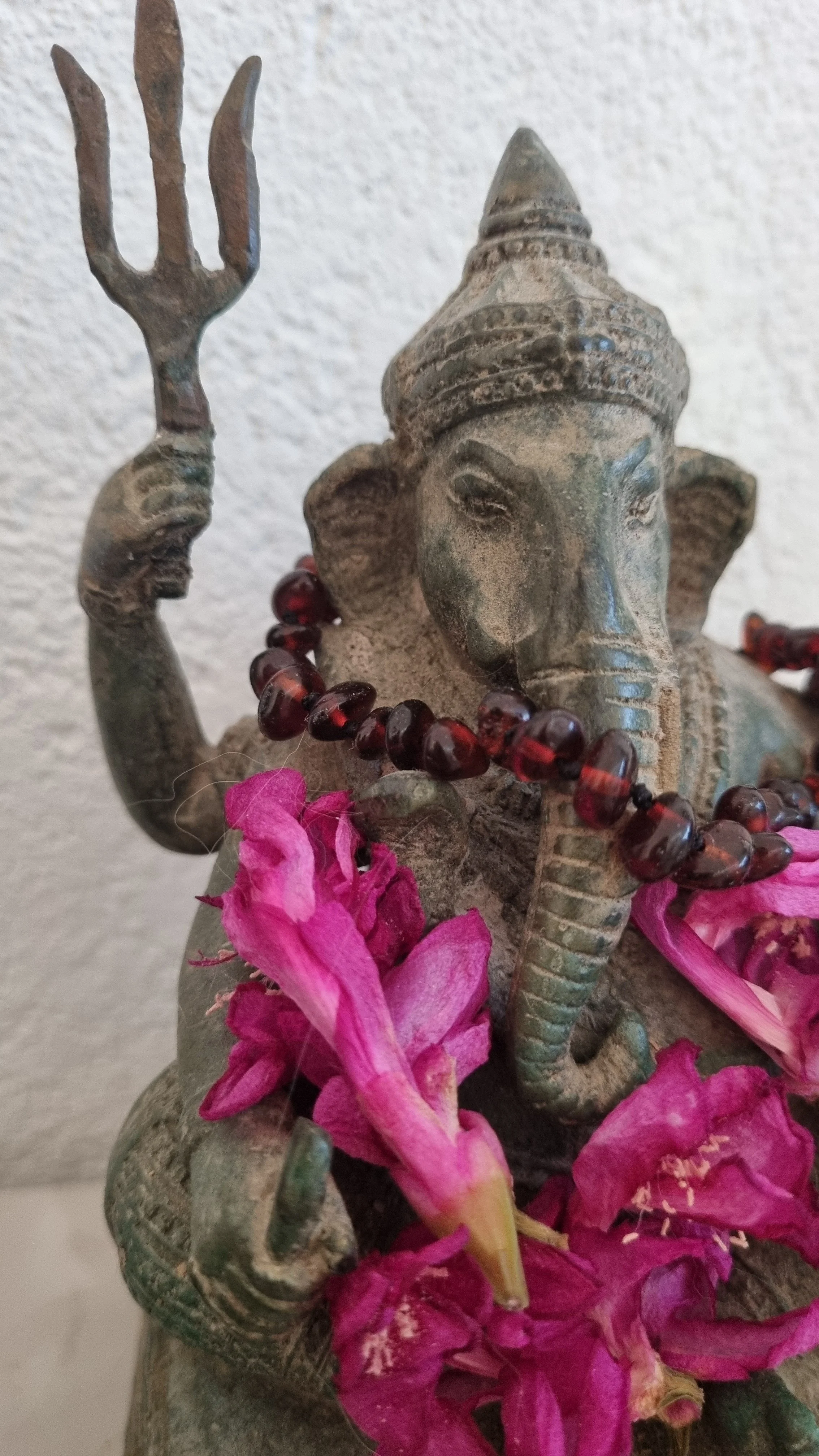 Eine geschnitzte Statue eines hinduistischen Gottes, vermutlich Ganesha, mit einer Halskette aus dunklen Perlen, pinken Blumen und einem großen spitzen Hut, gegen eine weiße Wand.