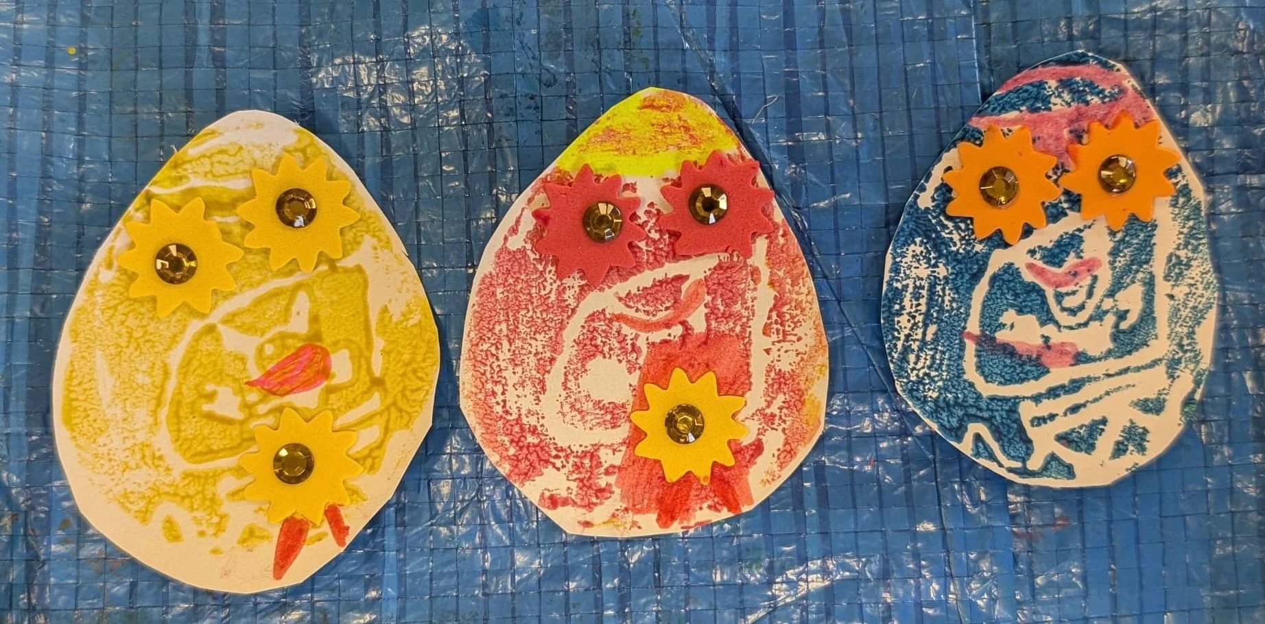 Three decorated paper eggs with yellow, red, and blue color designs, each adorned with small yellow and rhinestone embellishments, placed on a blue textured background.