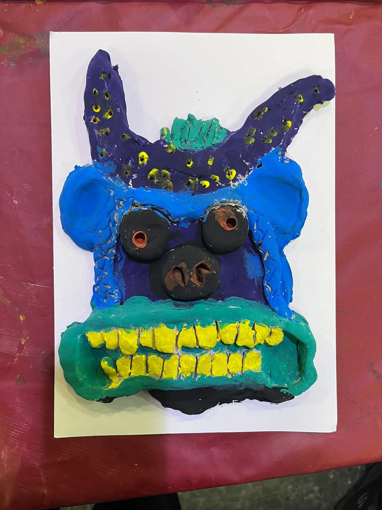 A colorful painted skull with exaggerated features, including large blue ears, a green and purple forehead, and prominent yellow teeth, on a white sheet of paper placed on a red table.