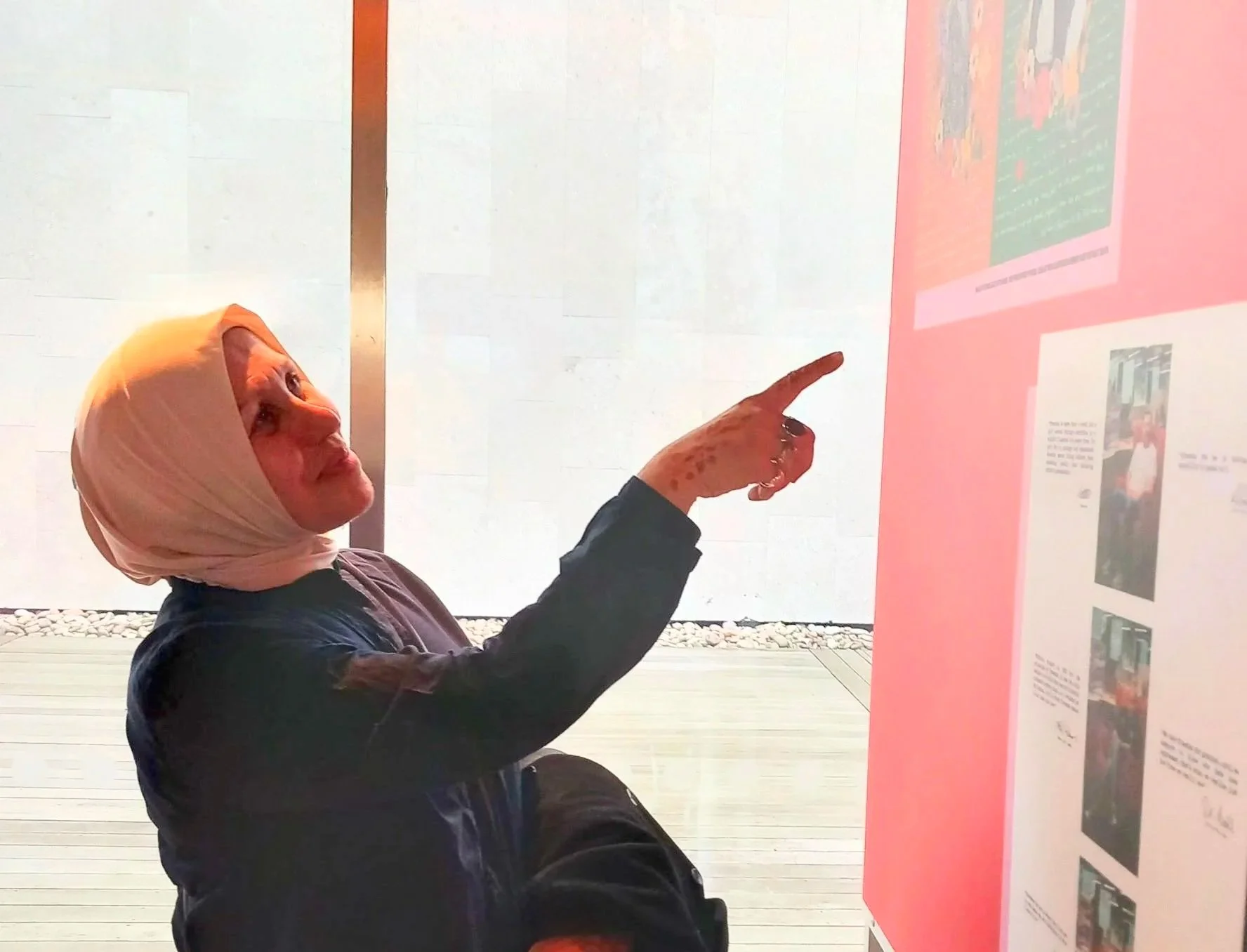 A woman with a light-colored headscarf pointing at a display board with photographs and text in an indoor setting.