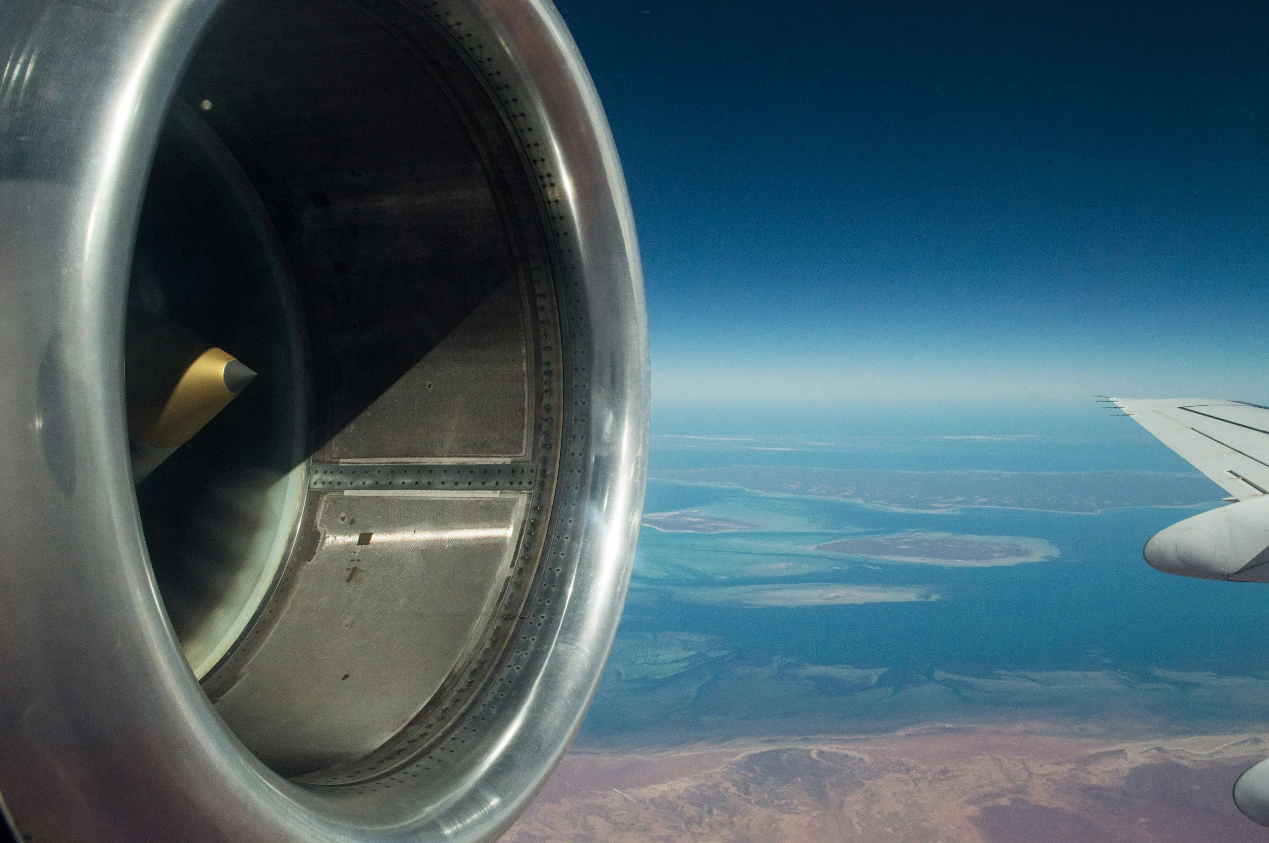 View from an airplane window showing a jet engine on the left and the wing on the right, with a landscape of lakes and land below and a clear blue sky.