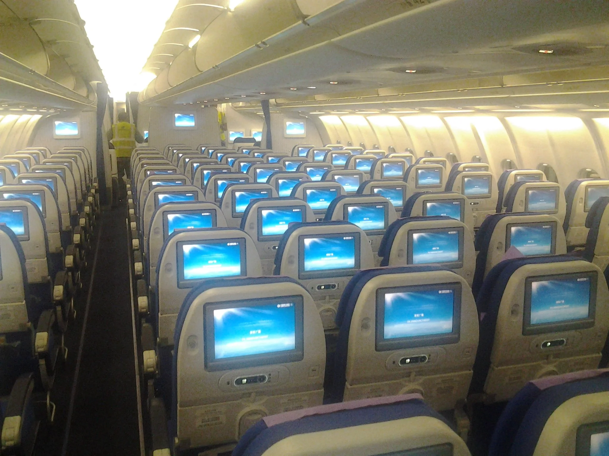 Inside an airplane cabin, showing rows of seats with personal entertainment screens on the back. There are overhead compartments and a few passengers, including a flight attendant in a yellow vest near the front.
