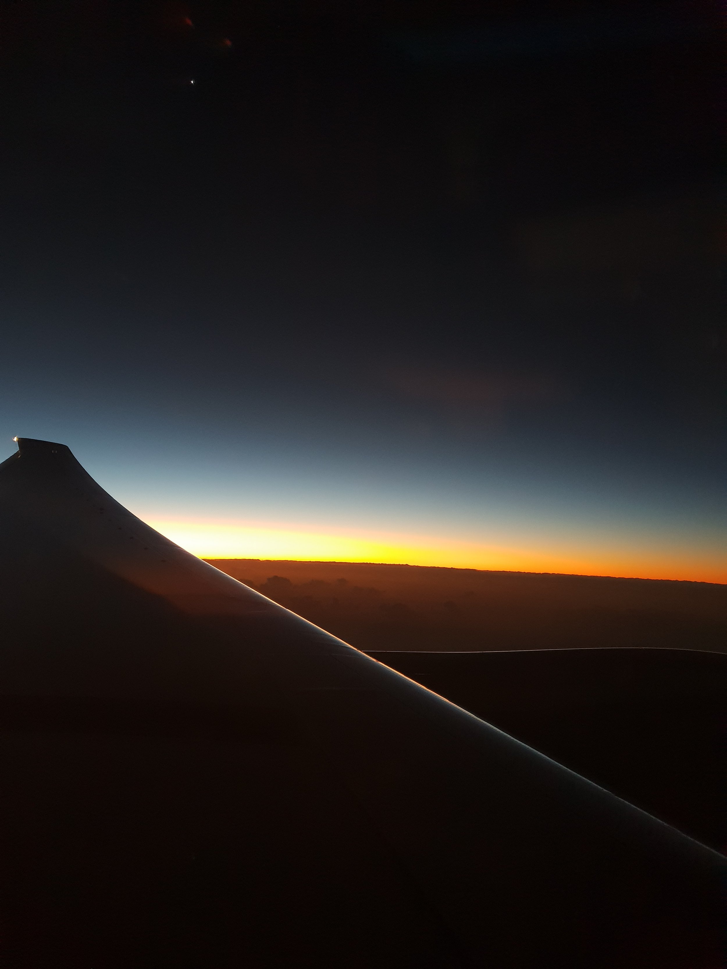 View from an airplane window showing a sunset or sunrise with the airplane wing in the foreground and a dark sky transitioning to bright horizon.
