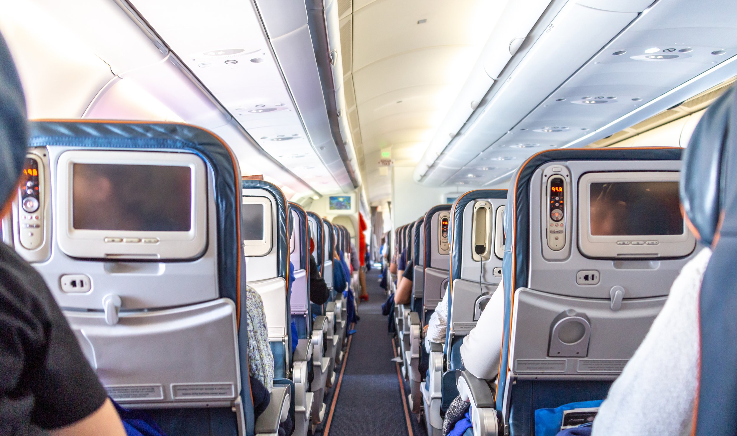Inside an airplane cabin with rows of seats and personal entertainment screens on the back of each seat, filled with passengers seated and looking forward.