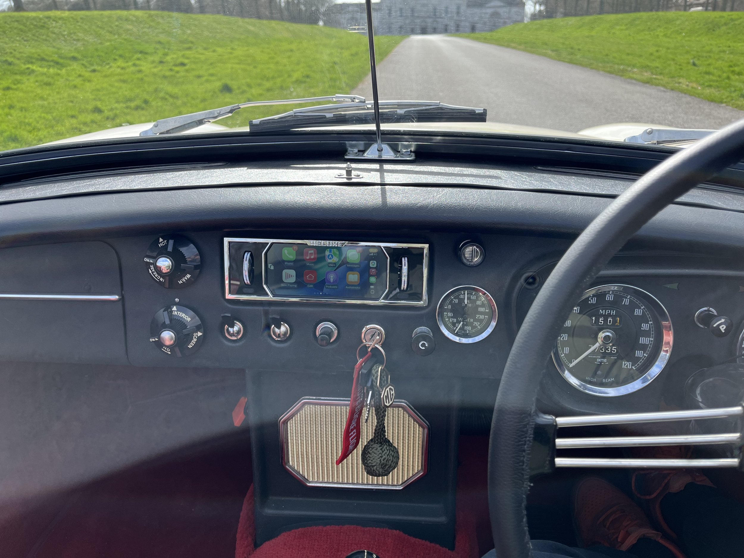 The dashboard and steering wheel of a vintage car on a rural road, with a smartphone mounted in the center console, and keys hanging from the ignition.