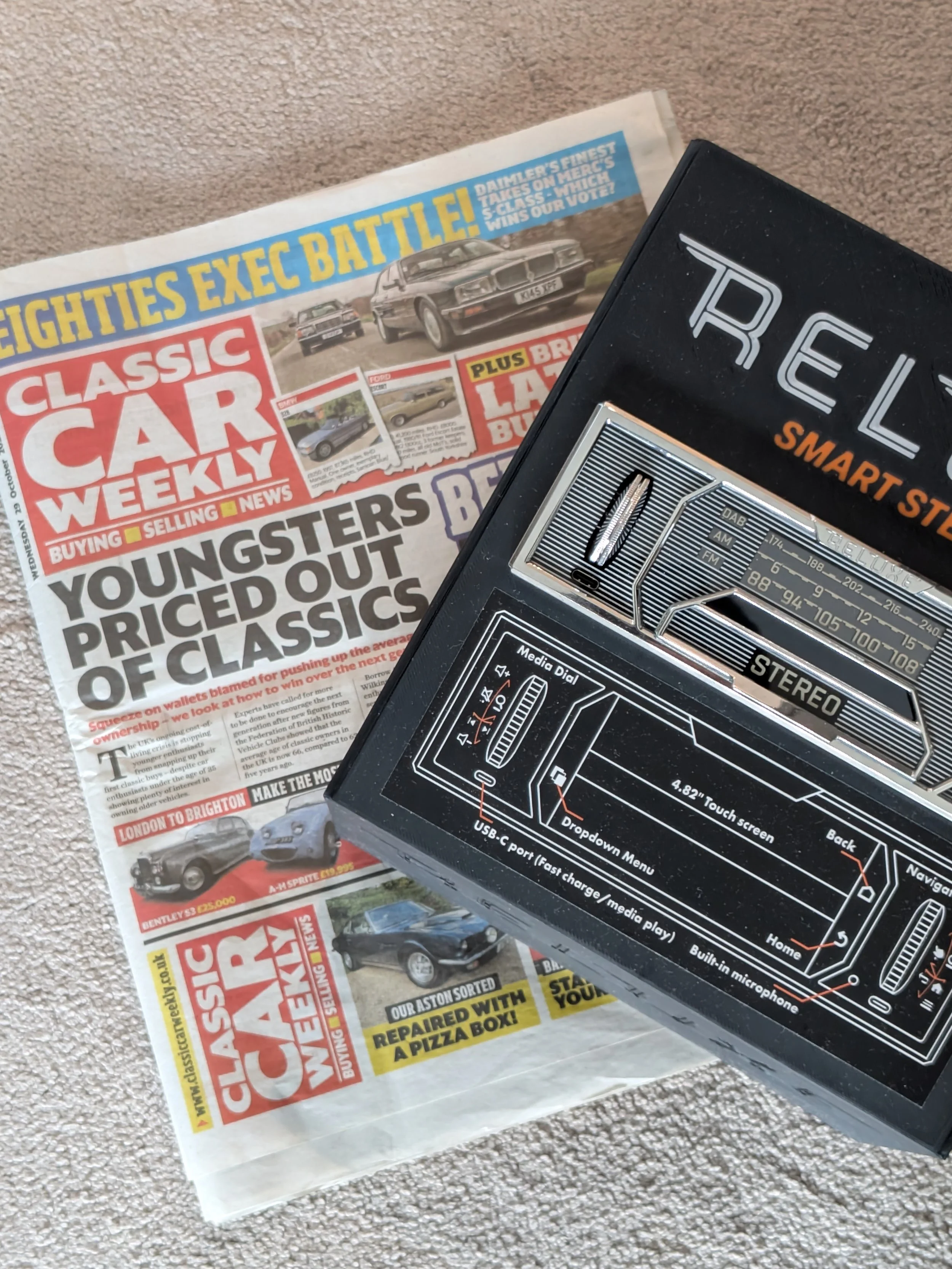 A newspaper titled 'Classic Car Weekly' with headlines about classic cars and an old-style transistor radio, both placed on a beige carpet.