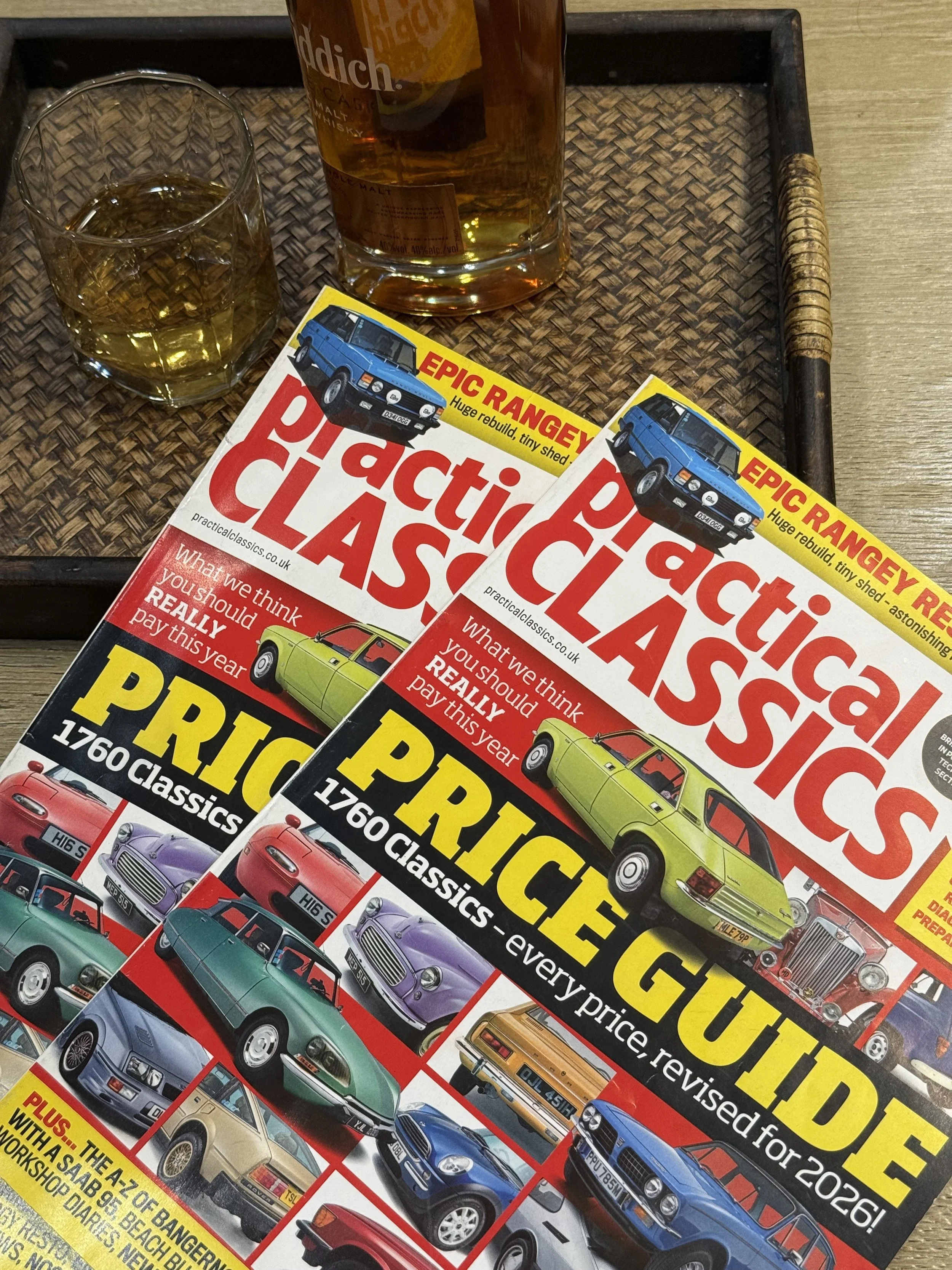 Two copies of 'Practical Classics' magazine on a wooden tray, with a glass of whiskey and a whiskey bottle in the background.