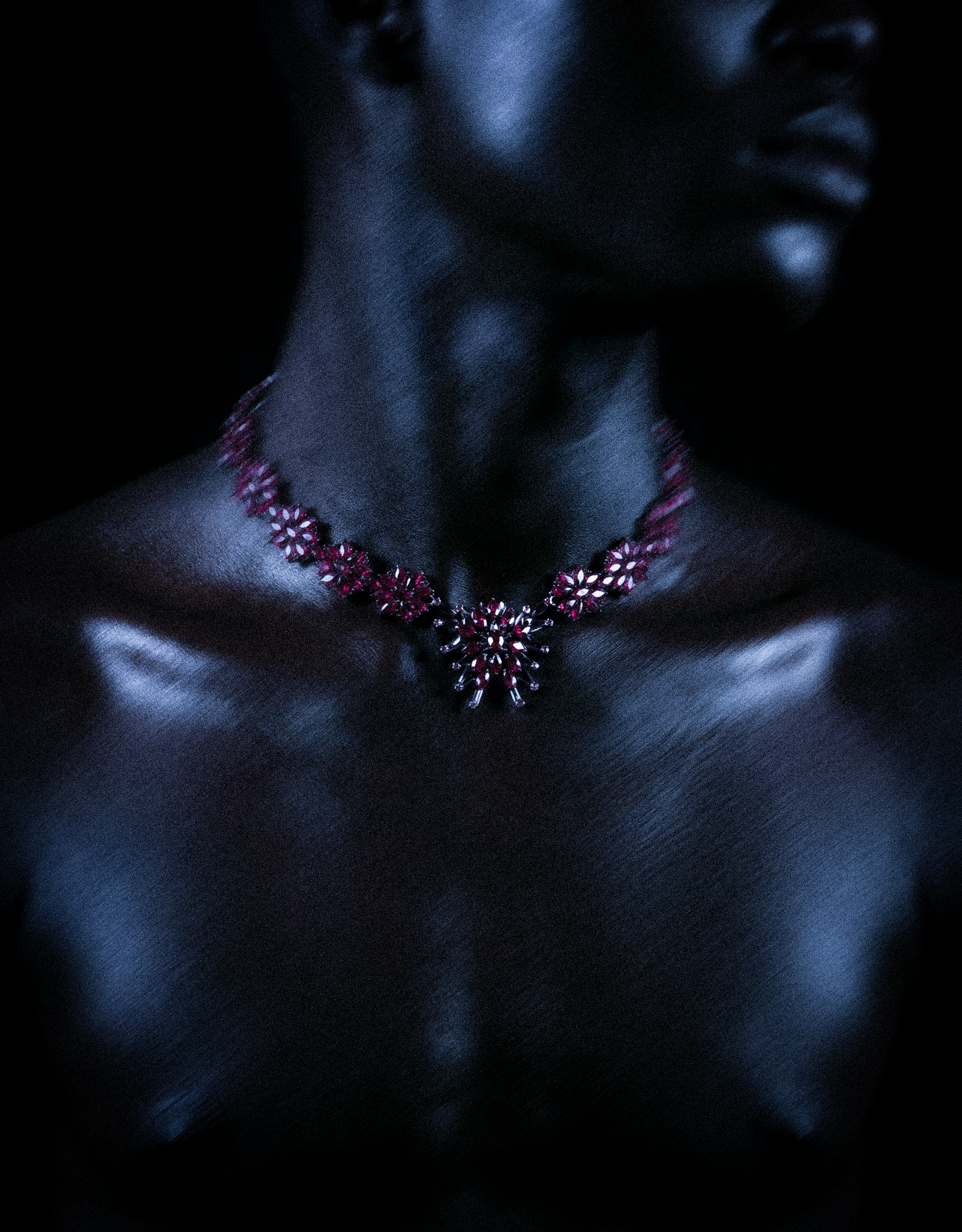 Close-up of a woman with dark skin wearing a red jewelry necklace, with a tattoo on her shoulder and a partial view of her face, against a black background.