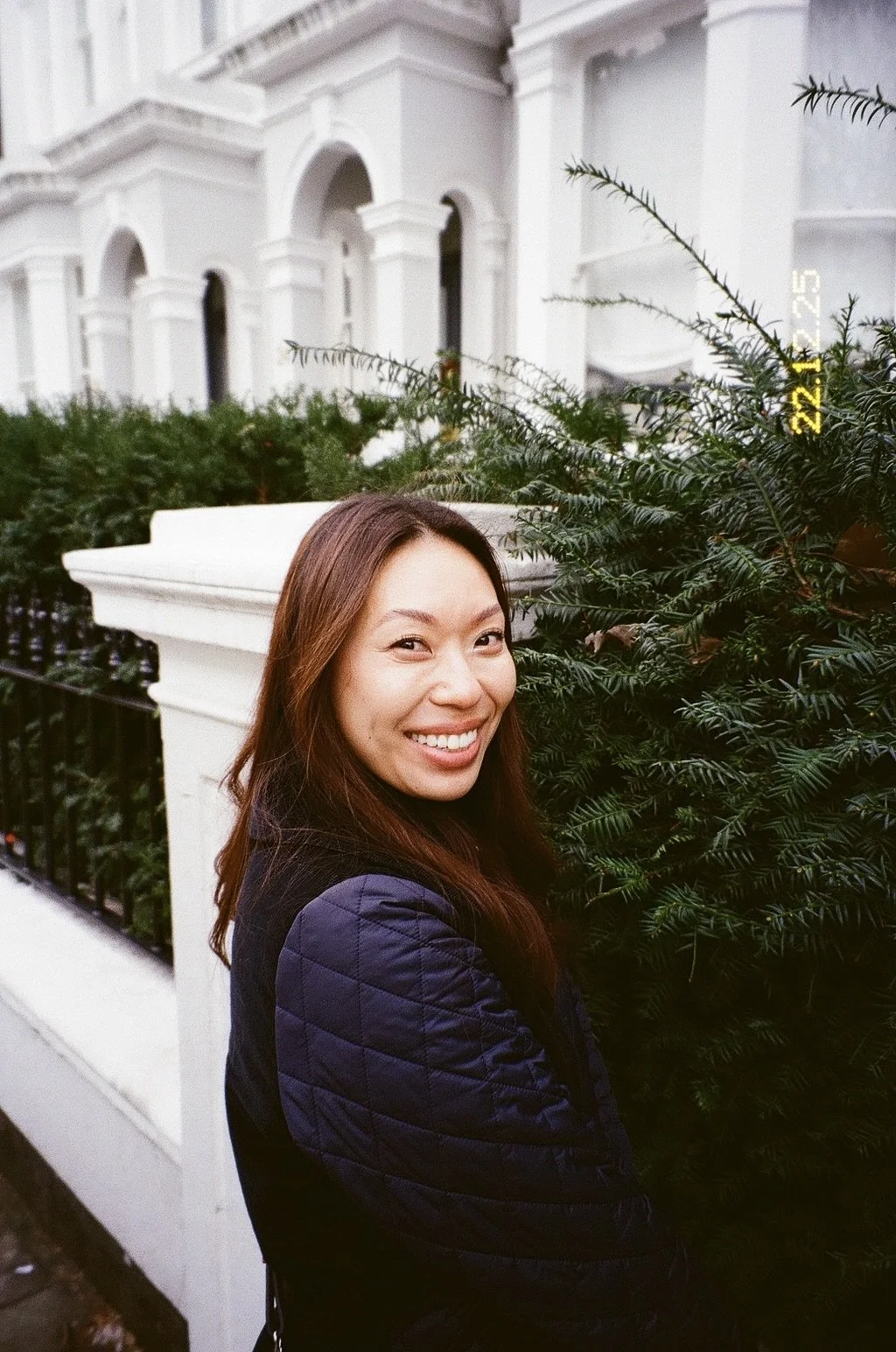 Smiling woman with long brown hair standing next to a bush in front of a white building with arched windows.