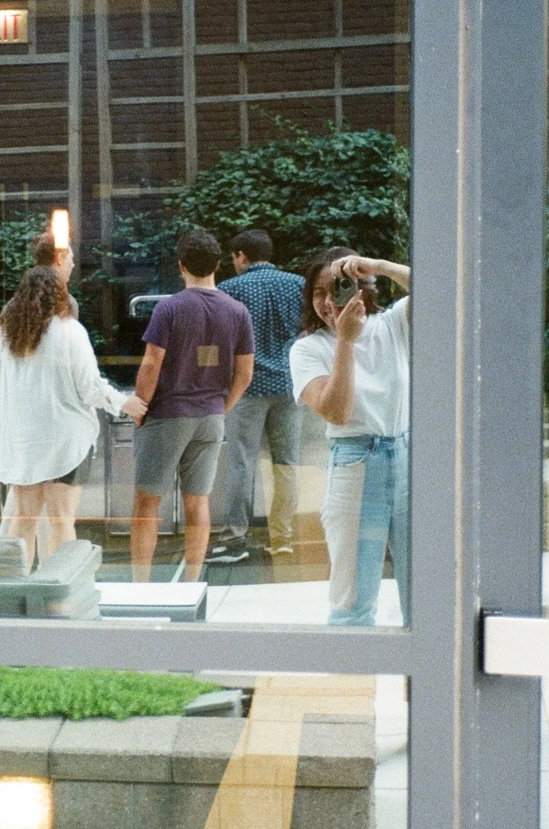 A woman in a white shirt and blue jeans taking a mirror selfie at a restaurant or cafe, with four people visible in the background, including two men and two women, one of whom is holding hands with the other.