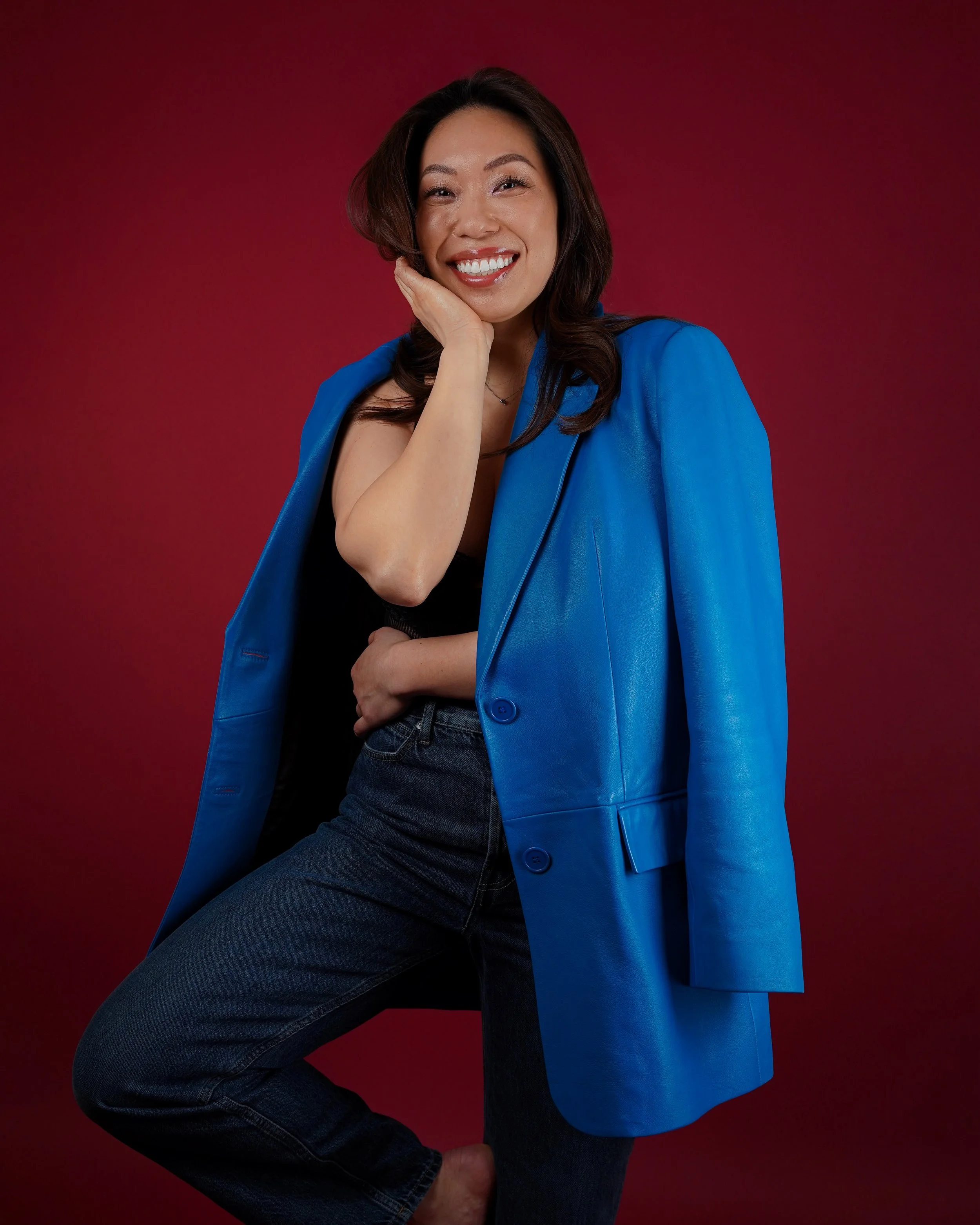 A smiling woman with dark hair, wearing a black top and a blue blazer draped over her shoulders, sitting on one knee against a red background.