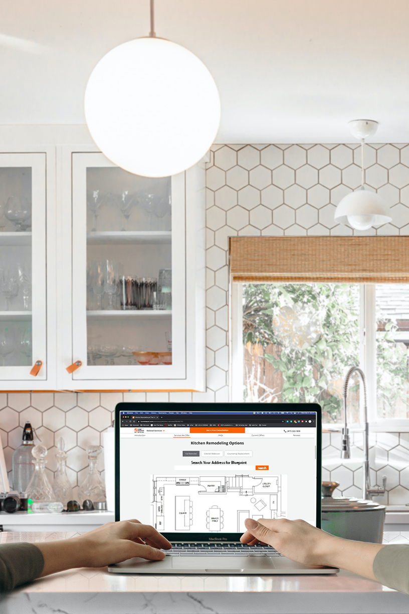 A person holding a laptop displaying a kitchen remodeling webpage in a bright kitchen with white cabinets, hexagonal tile backsplash, and a window with a bamboo shade.