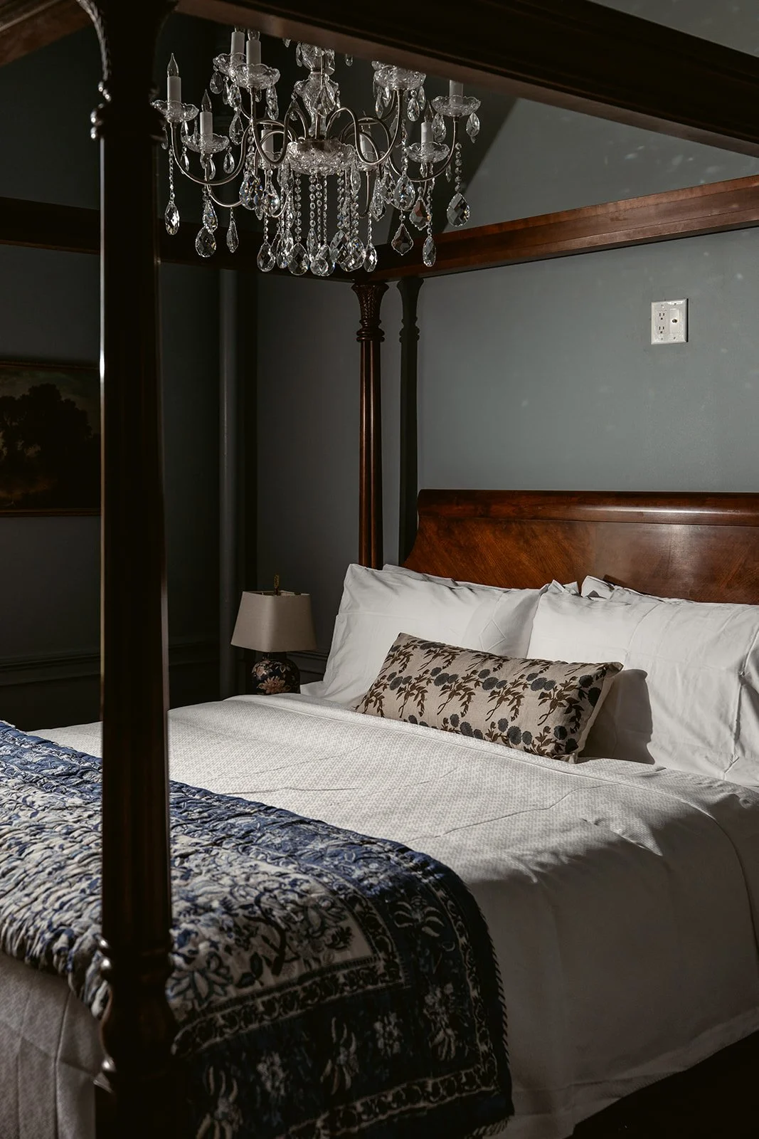 A bedroom with a chandelier hanging from a dark ceiling, a wooden four-poster bed with white bedding and decorative pillows, a bedside table with a lamp, and a dark painted wall. Hotel Everett | Boutique Hotel in Winnsboro, SC, historic hotel