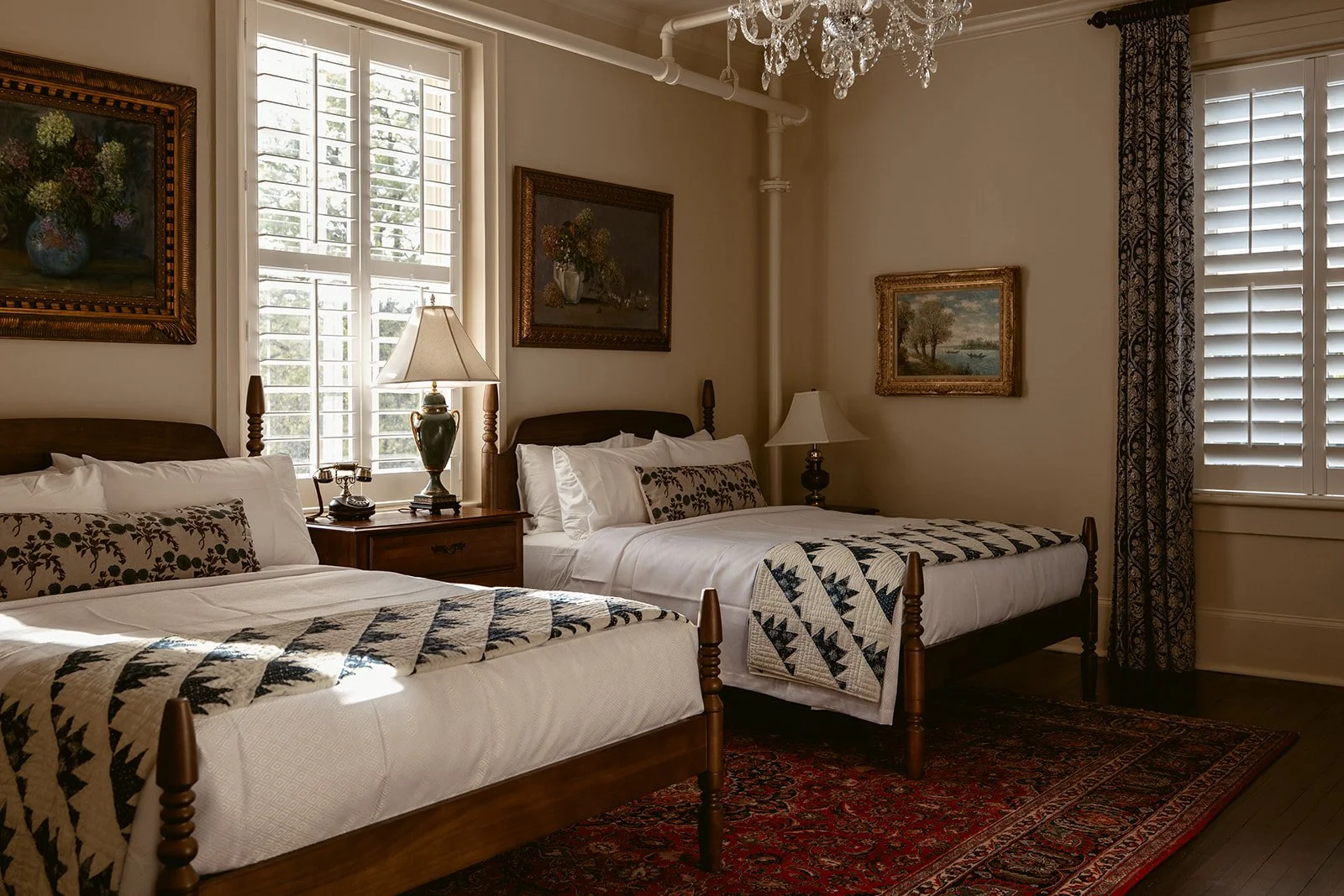 A cozy bedroom with two twin beds with wooden frames, white bedding, and decorative pillows. There are two windows with white shutters and patterned curtains, and a chandelier hanging from the ceiling. The room has framed paintings on the walls, two bedside tables with lamps, and a vintage rug on the floor.