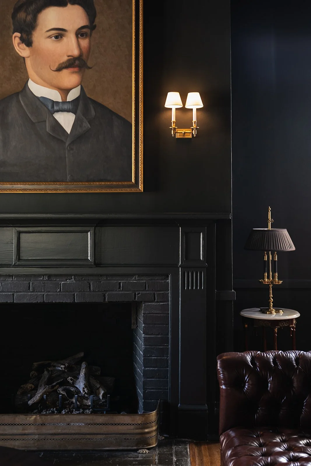 Interior of a room with a black fireplace,sitting below a large portrait of a man with a mustache and wearing a dark suit.  To the right, a wall-mounted lamp with two shades and a table lamp with a dark shade and gold accents are visible. There is a brown leather armchair in the lower right corner.