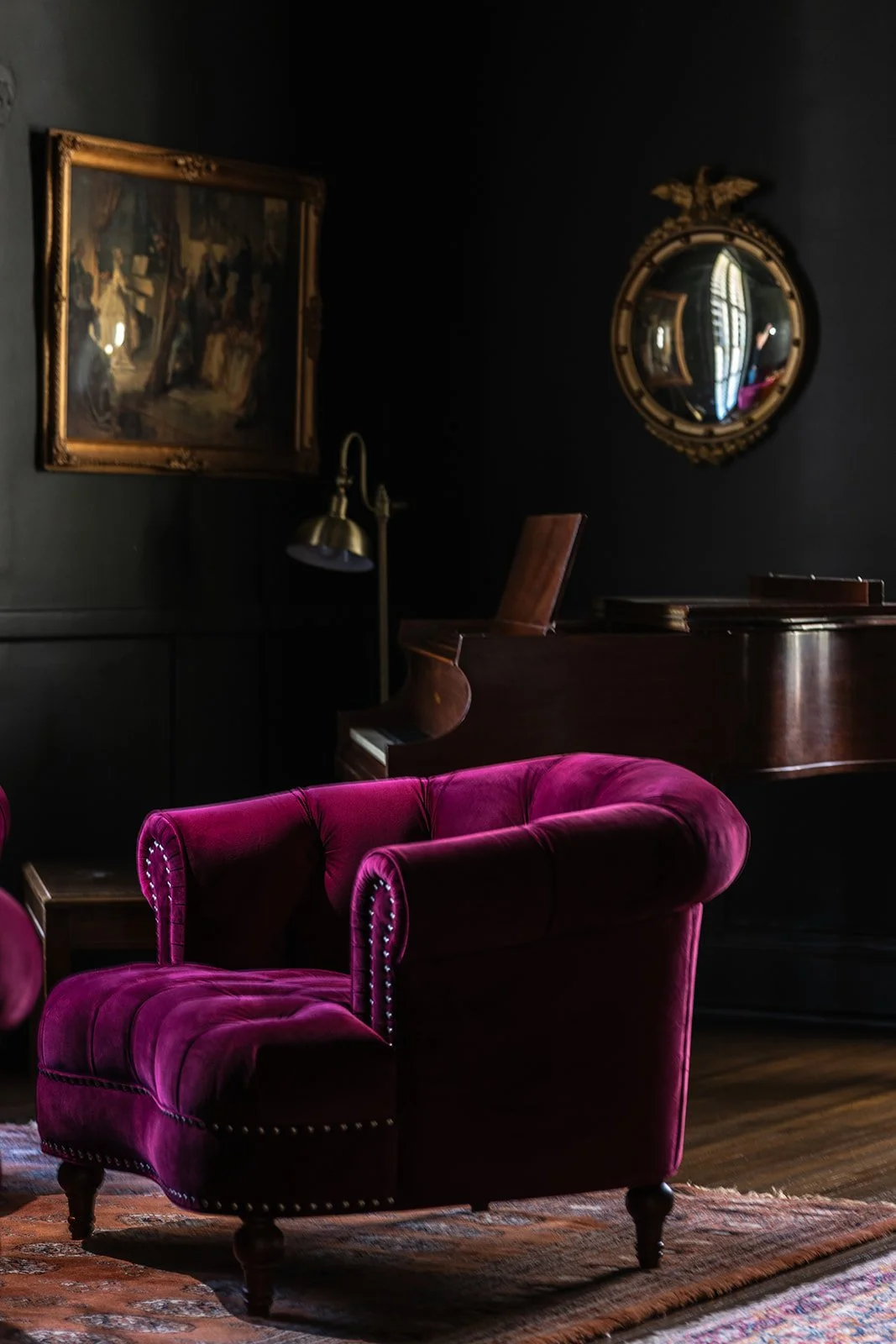 A vintage styled room with dark walls, featuring a purple velvet tufted armchair, a wall mirror with an ornate gold frame, a painting, a polished wooden piano, and a brass wall lamp.