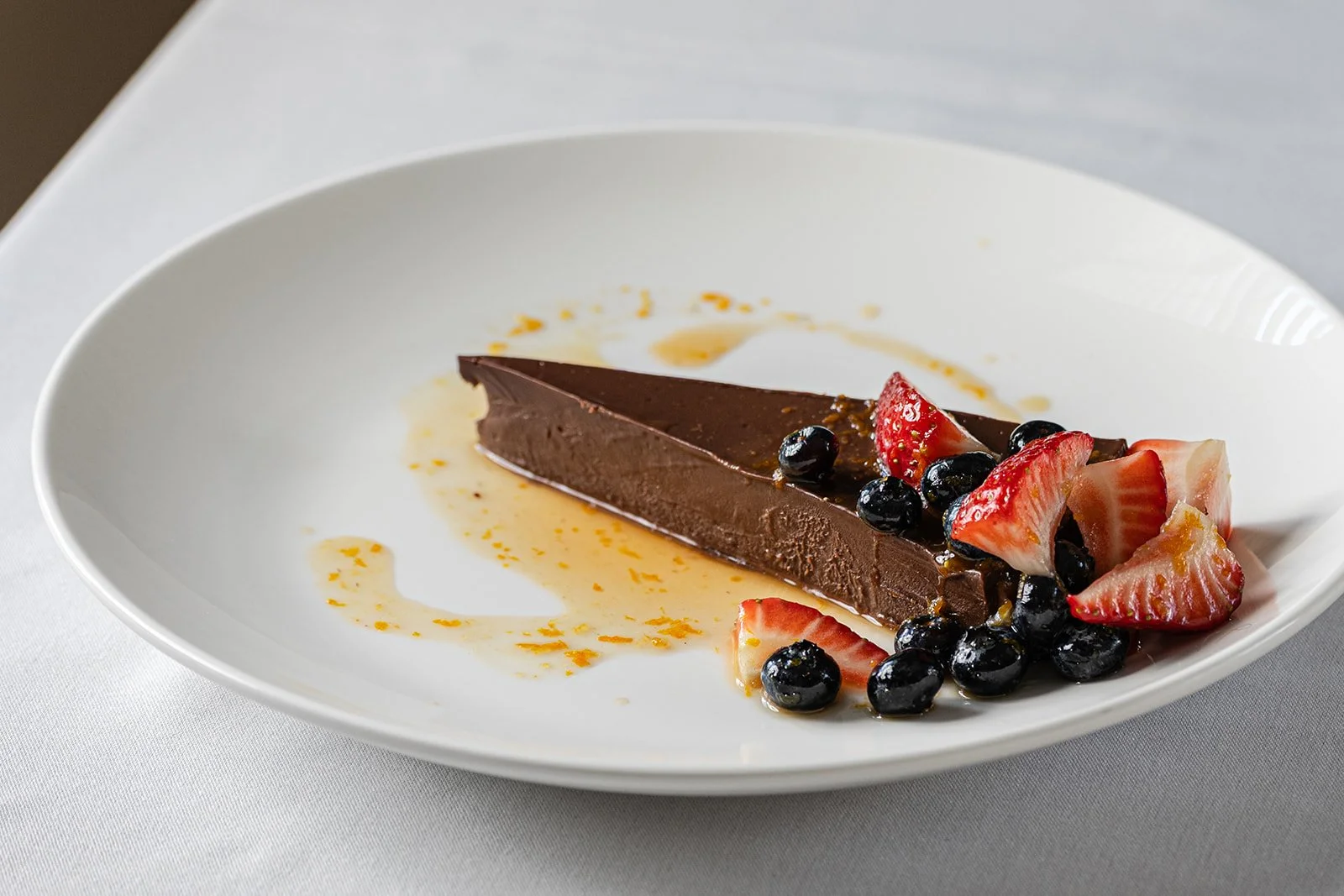 A slice of chocolate cake garnished with strawberries and blueberries, on a white plate with some sauce or syrup drizzled around.