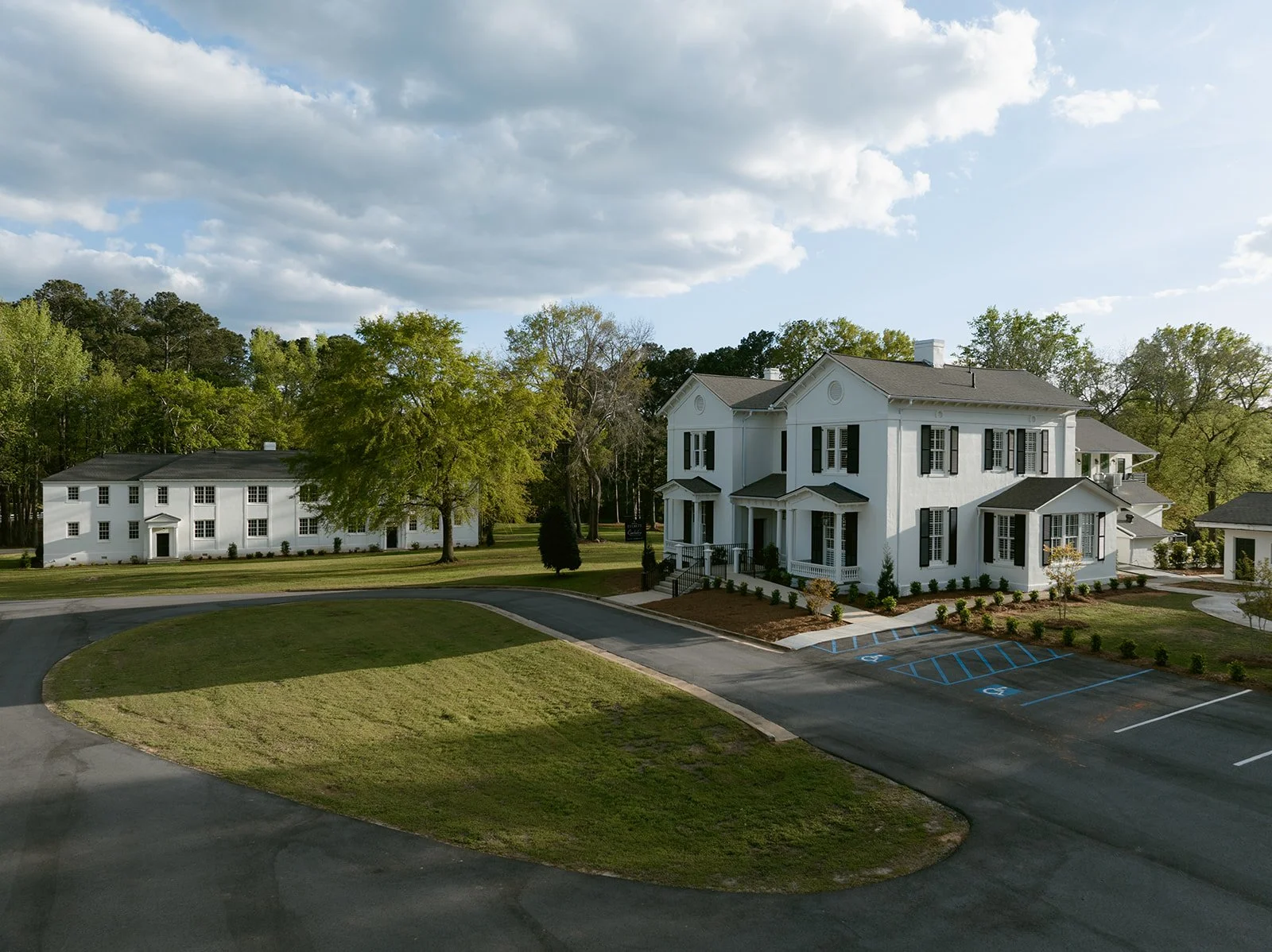 Large white two-story house with black shutters, a porch, and a driveway with parking for handicapped. Smaller white building in the background, surrounded by trees and green lawns under a partly cloudy sky.