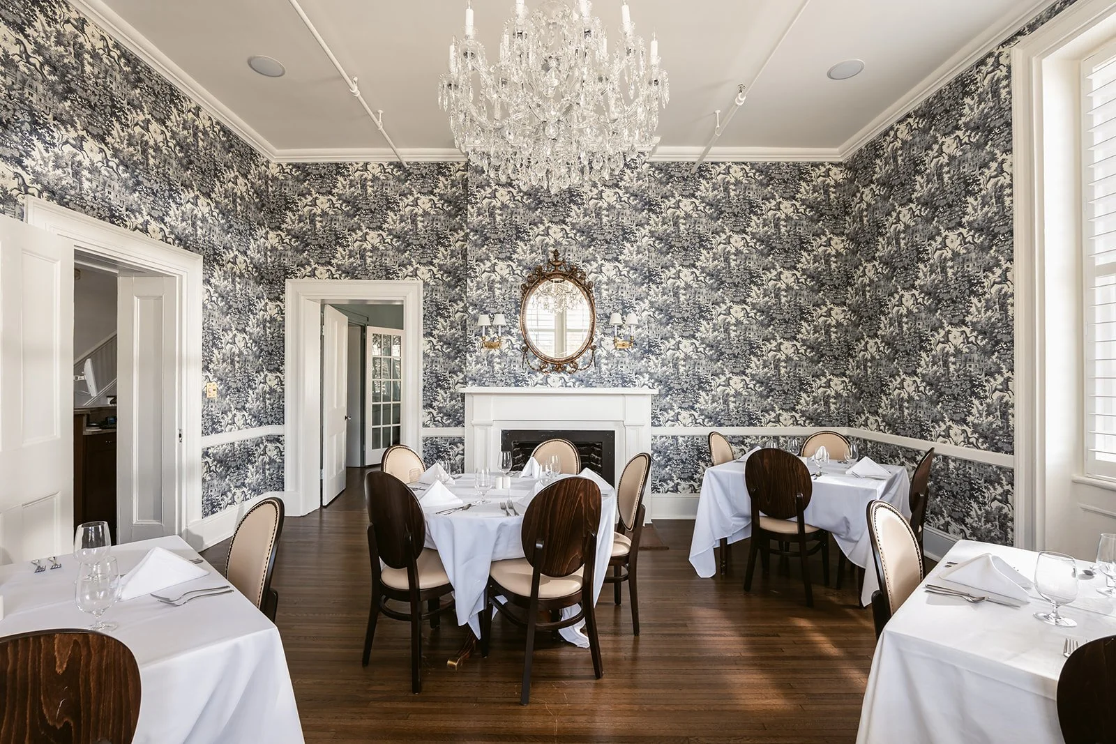 Elegant dining room with white tablecloths, napkins, wine glasses, and cutlery, decorated with a crystal chandelier, ornate mirror, and patterned wallpaper.